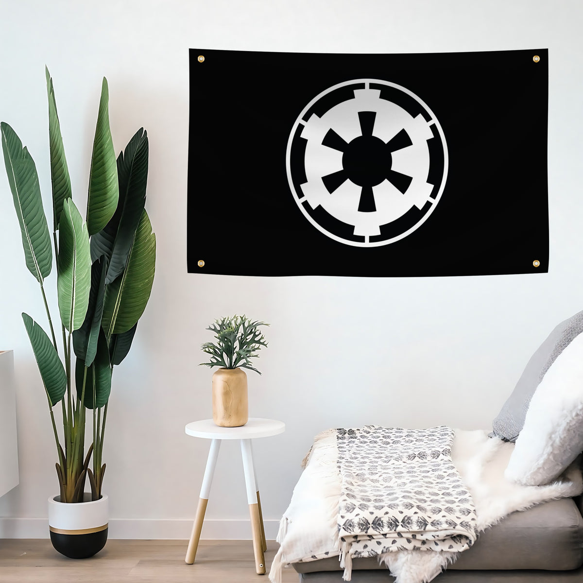 A black wall flag banner featuring the white Star Wars Galactic Empire crest, hanging on a white wall in a styled room setting with a plant, side table, and sofa.