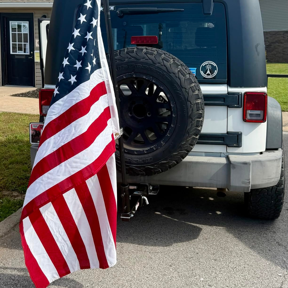 Back of a jeep wrangler with an American flag attached to the hitch, parked in a driveway.