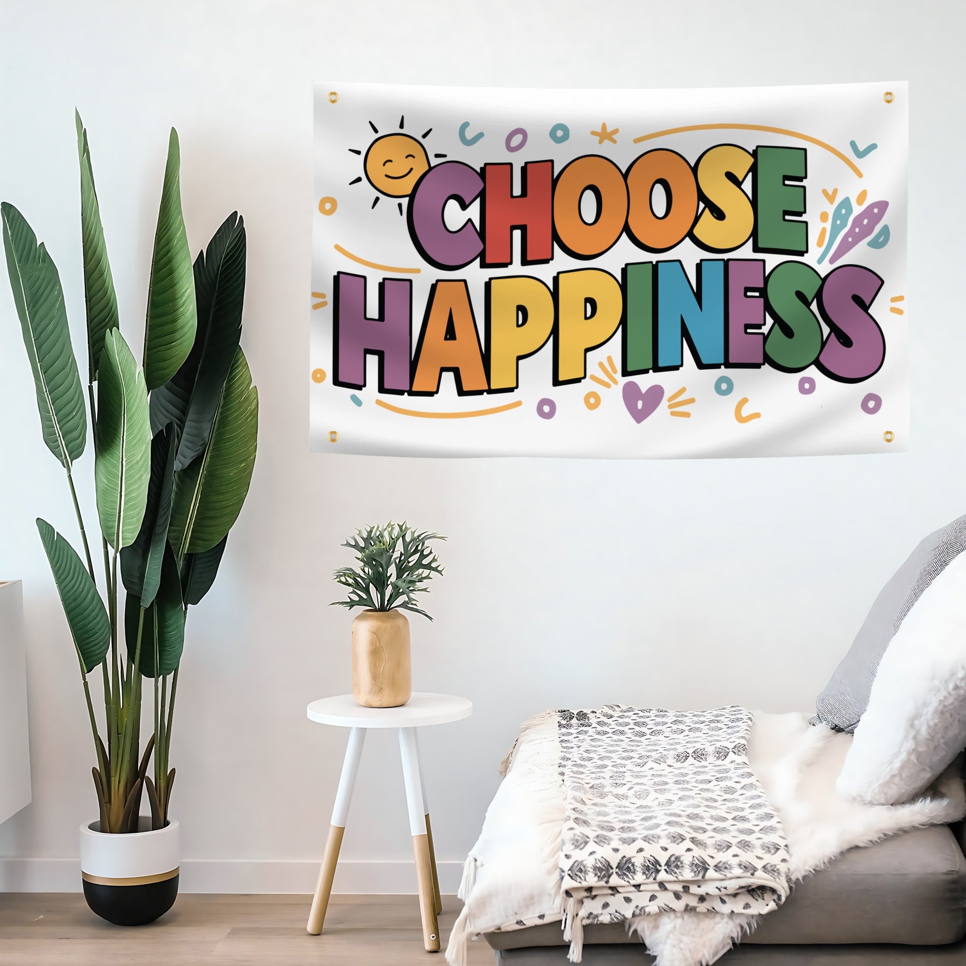 A colorful flag banner featuring the text 'CHOOSE HAPPINESS' in playful multicolor bubble letters, hanging on a white wall in a styled living room setting with a plant, side table, and sofa.