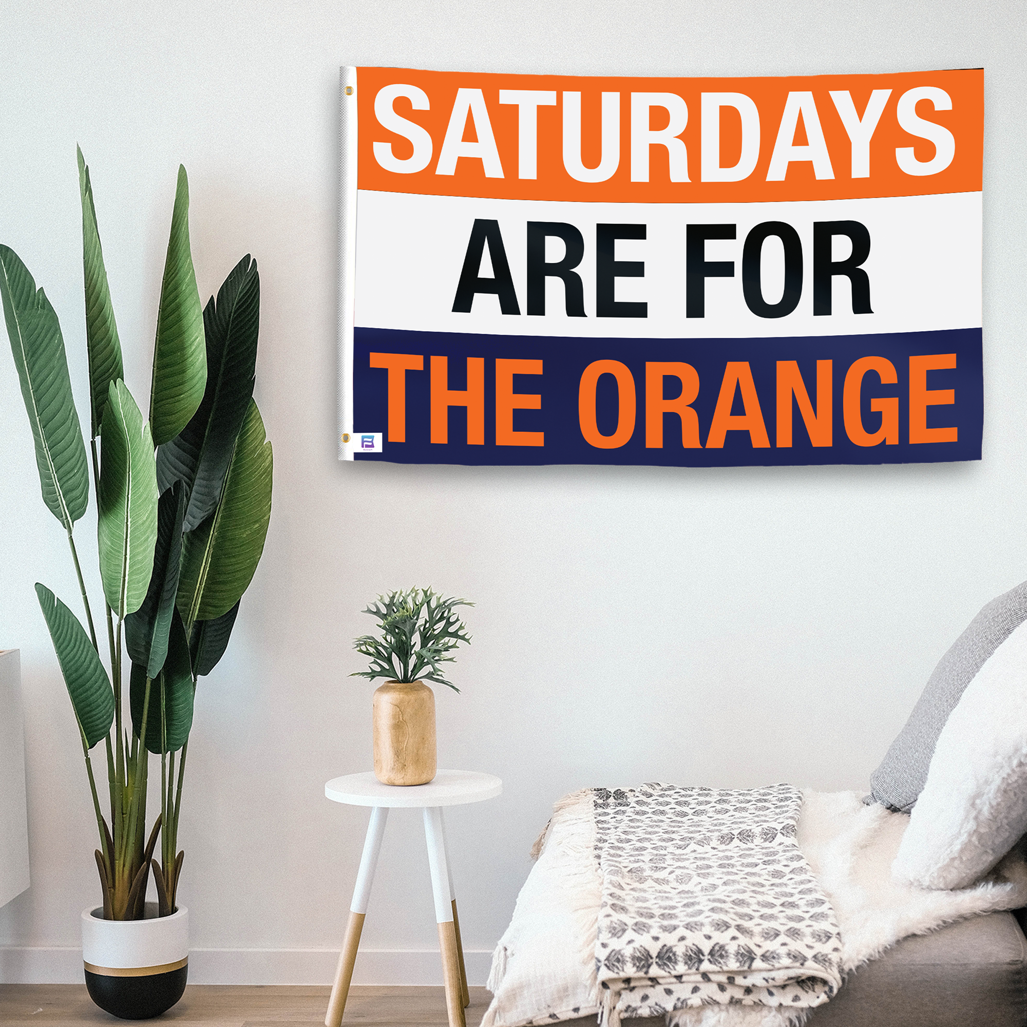 In a home setting, a flag with the saying "Saturdays Are for the Orange" is mounted on a white wall by a side table.