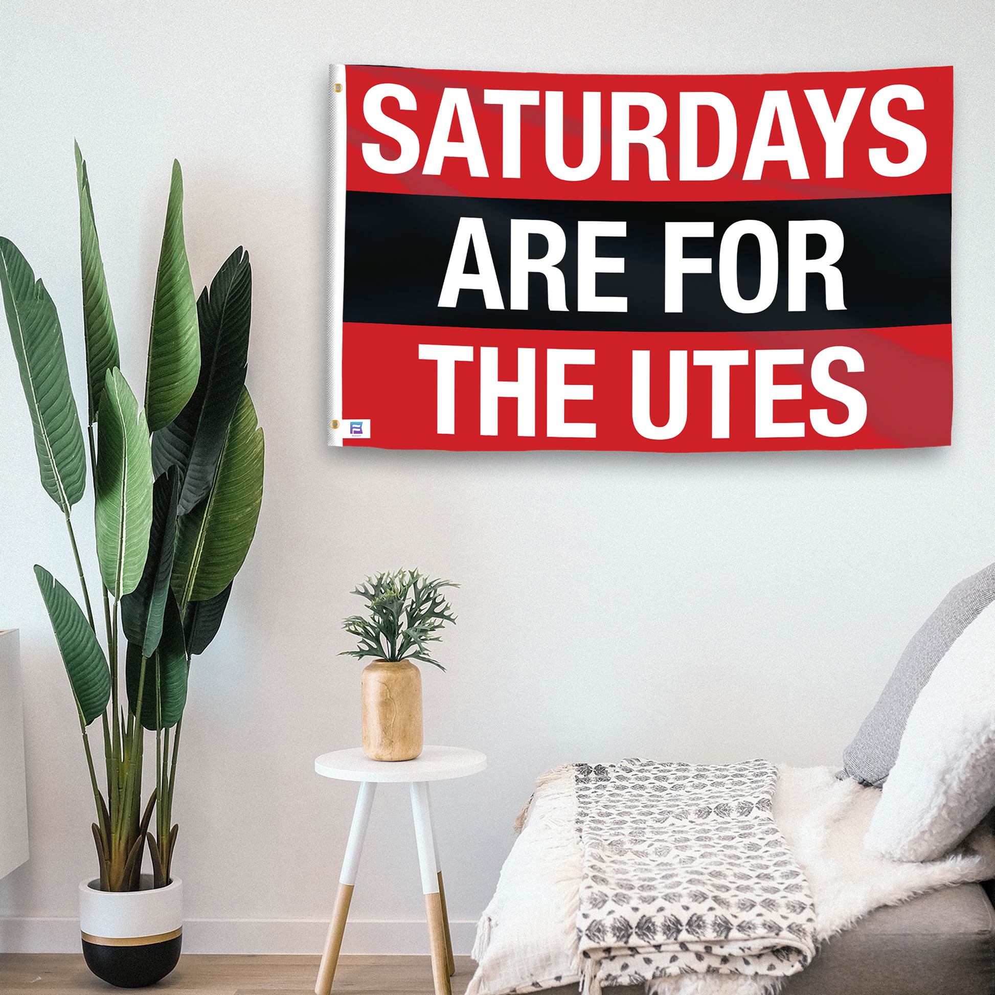 In a home setting, a flag with the saying "Saturdays Are for the Utes" is mounted on a white wall by a side table.