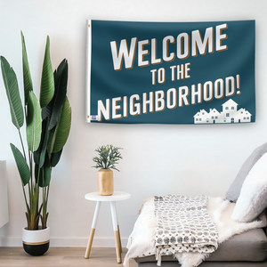 In a home setting, a flag with the saying "Welcome To The Neighborhood" is mounted on a white wall by a side table.