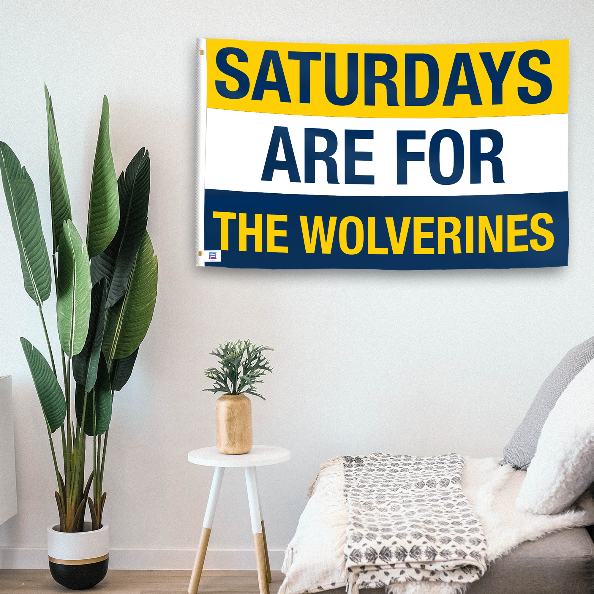 In a home setting, a flag with the saying "Saturdays Are for the Wolverines" is mounted on a white wall by a side table.