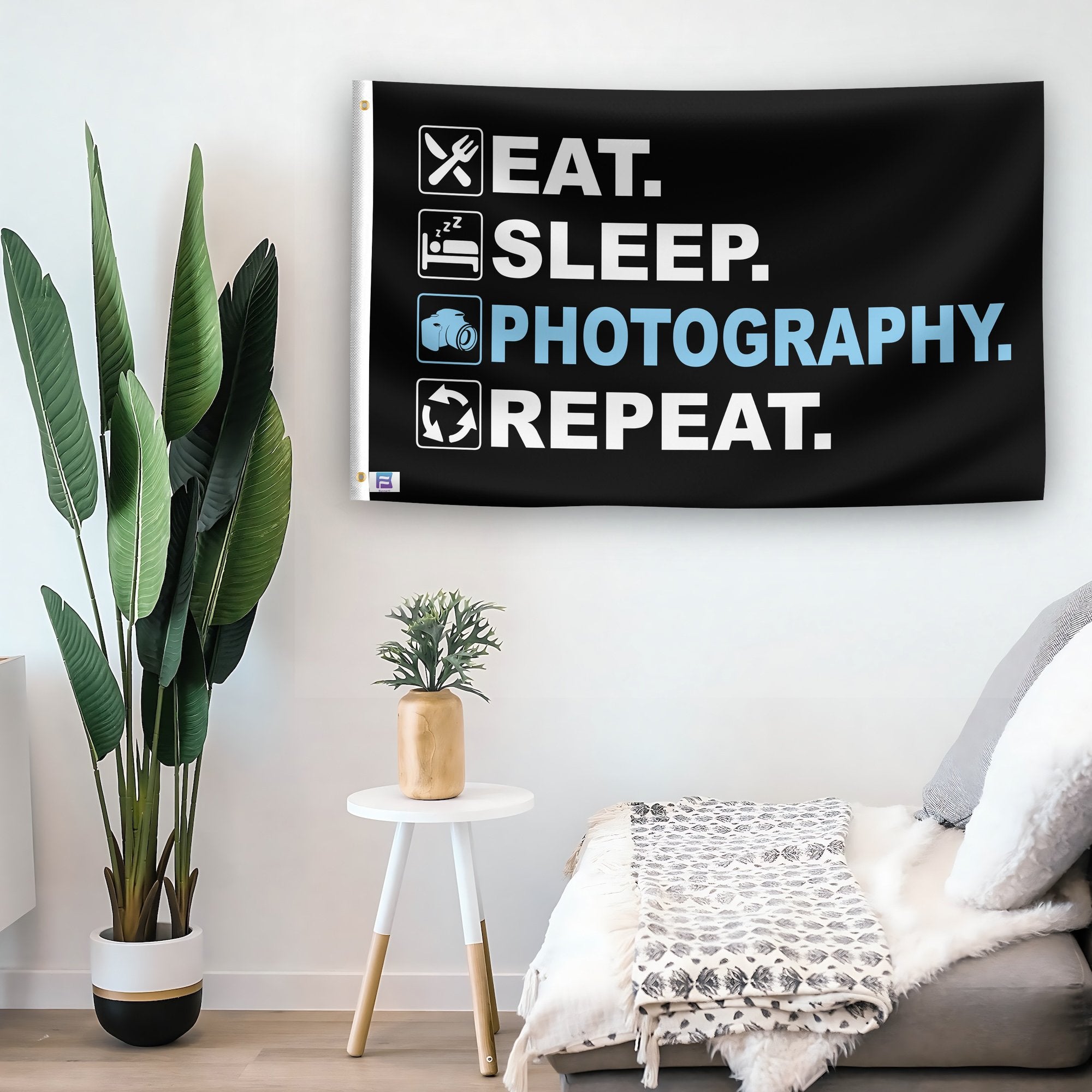 In a home setting, a flag with the saying "Eat Sleep Photography Repeat" is mounted on a white wall by a side table.