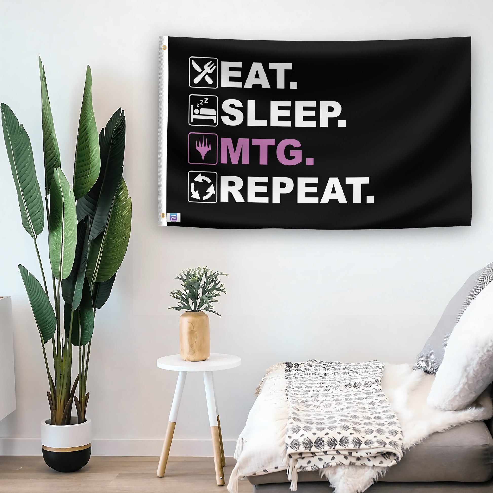 In a home setting, a flag with the saying "Eat Sleep MTG Repeat" is mounted on a white wall by a side table.