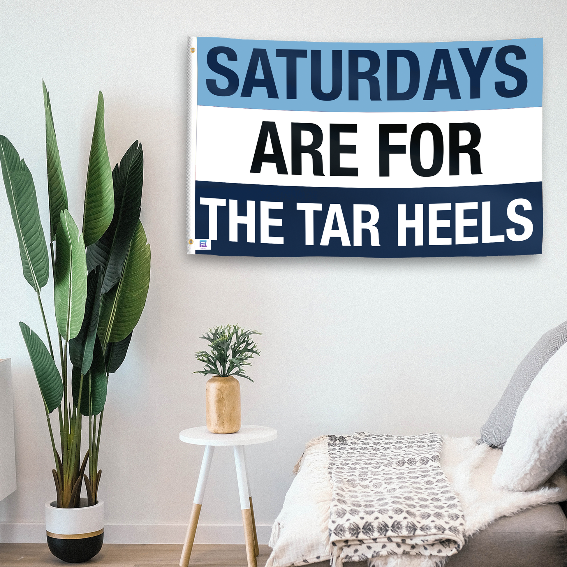 In a home setting, a flag with the saying "Saturdays Are for the Tar Heels" is mounted on a white wall by a side table.