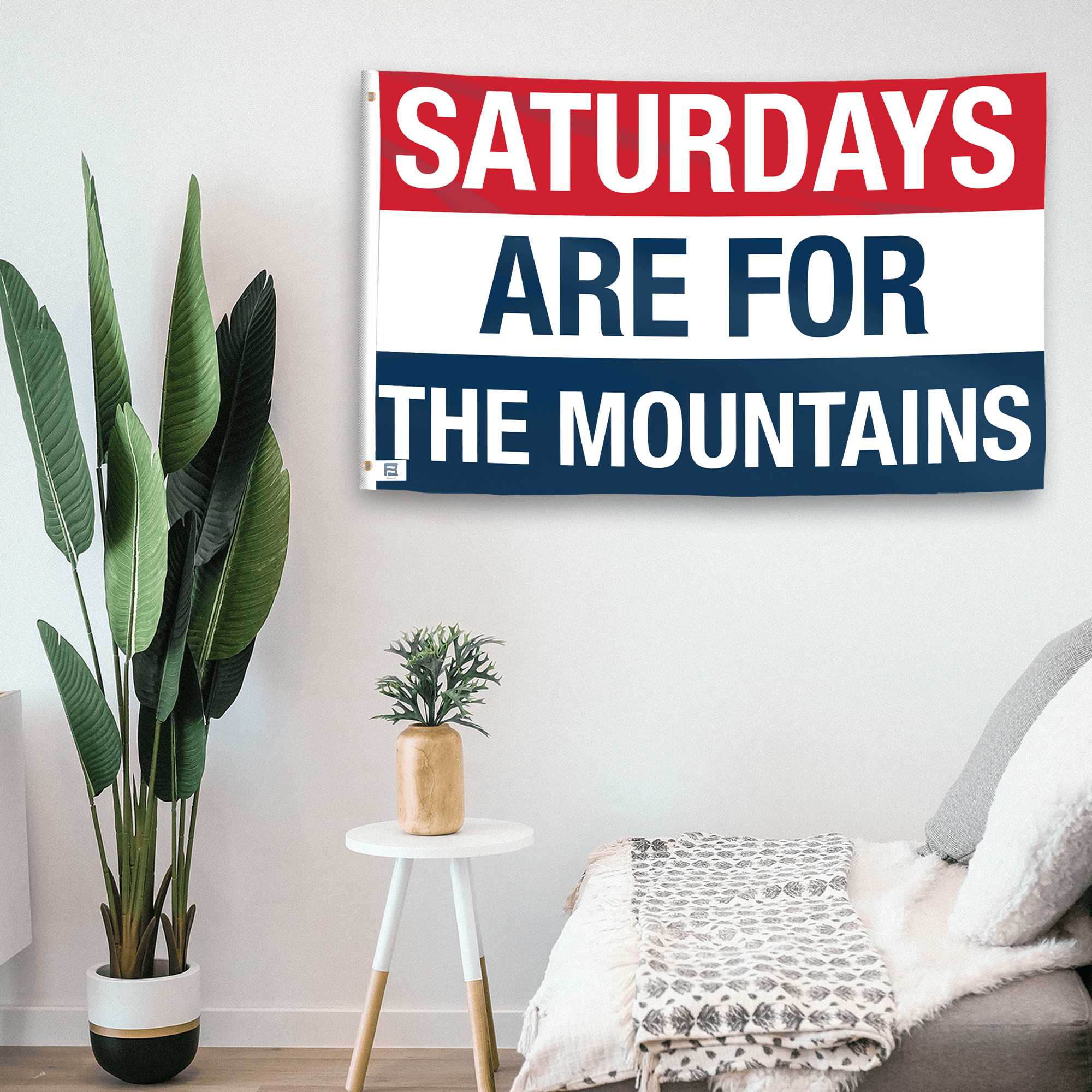 In a home setting, a flag with the saying "https://www.dropbox.com/scl/fi/vjm7ribm8e3ya9g0q947q/saturdays-are-for-the-mountains_room.png?rlkey=pdy15xzn72jj4ji3xx0o8o5vz&raw=1" is mounted on a white wall by a side table.