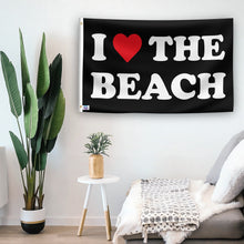 Load image into Gallery viewer, In a home setting, a flag with the saying &quot;I Love The Beach&quot; is mounted on a white wall by a side table.
