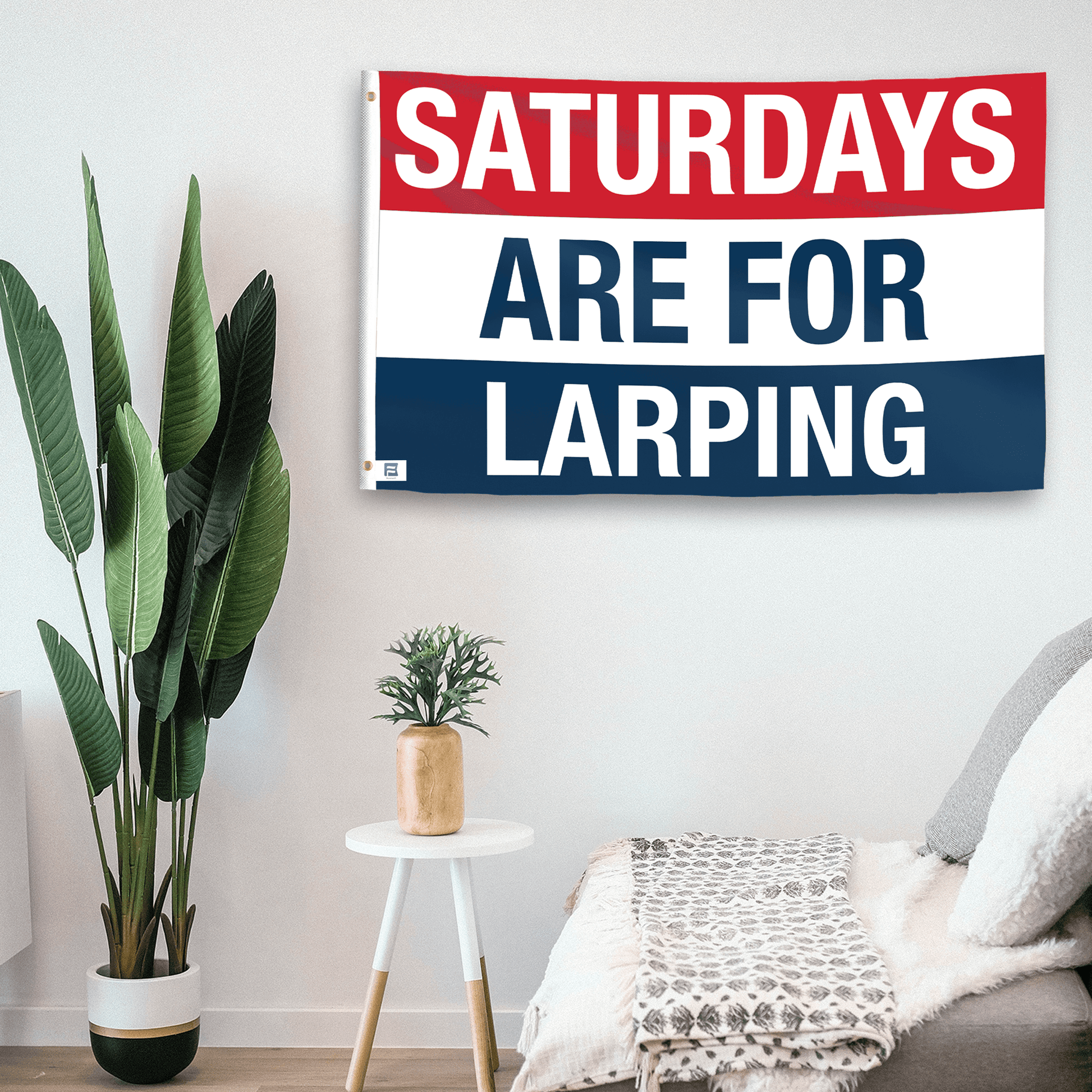 In a home setting, a flag with the saying "https://www.dropbox.com/scl/fi/65usy8v6uyey1g1tt9wp1/saturdays-are-for-larping_room.png?rlkey=m55txjhhx53jani4fldinw5fd&raw=1" is mounted on a white wall by a side table.