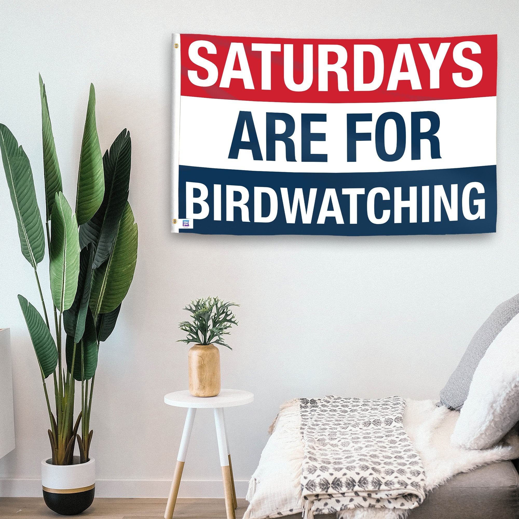 In a home setting, a flag with the saying "https://www.dropbox.com/scl/fi/mxuh2woaitq81khwlf9u6/saturdays-are-for-birdwatching_room.png?rlkey=liqm00sfkbnlrmsbsr1217tsx&raw=1" is mounted on a white wall by a side table.