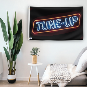 In a home setting, a flag with the saying "Tune Up" is mounted on a white wall by a side table.