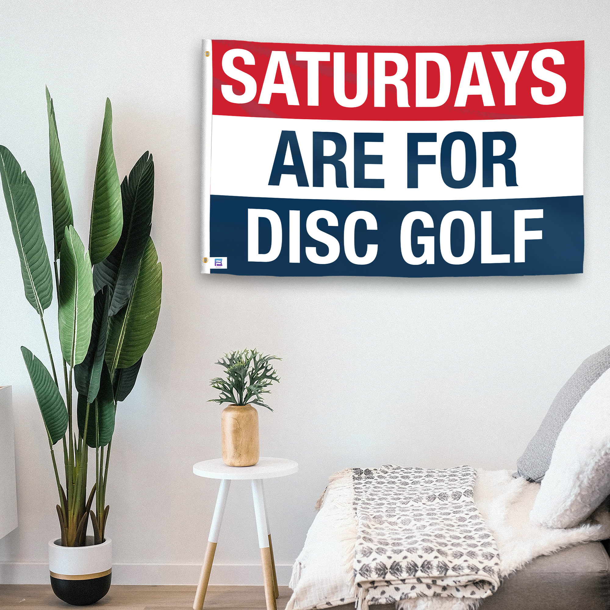 In a home setting, a flag with the saying "https://www.dropbox.com/scl/fi/jx4bk1noz36l78yeb95uh/saturdays-are-for-disc-golf_room.png?rlkey=l08scfv6tqo31ln54x2j6k44l&raw=1" is mounted on a white wall by a side table.