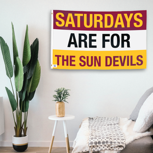 In a home setting, a flag with the saying "Saturdays Are for the Sun Devils" is mounted on a white wall by a side table.