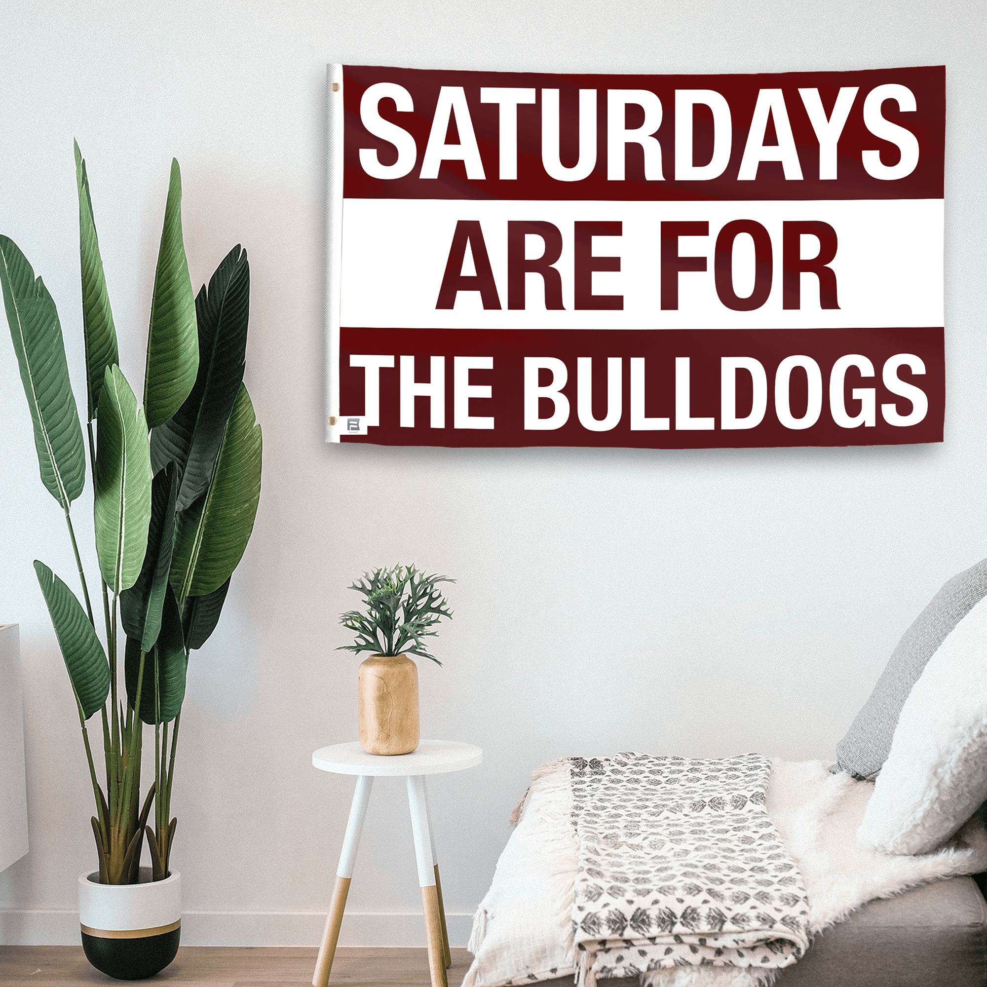 In a home setting, a flag with the saying "Saturdays Are for the Bulldogs Mississippi" is mounted on a white wall by a side table.