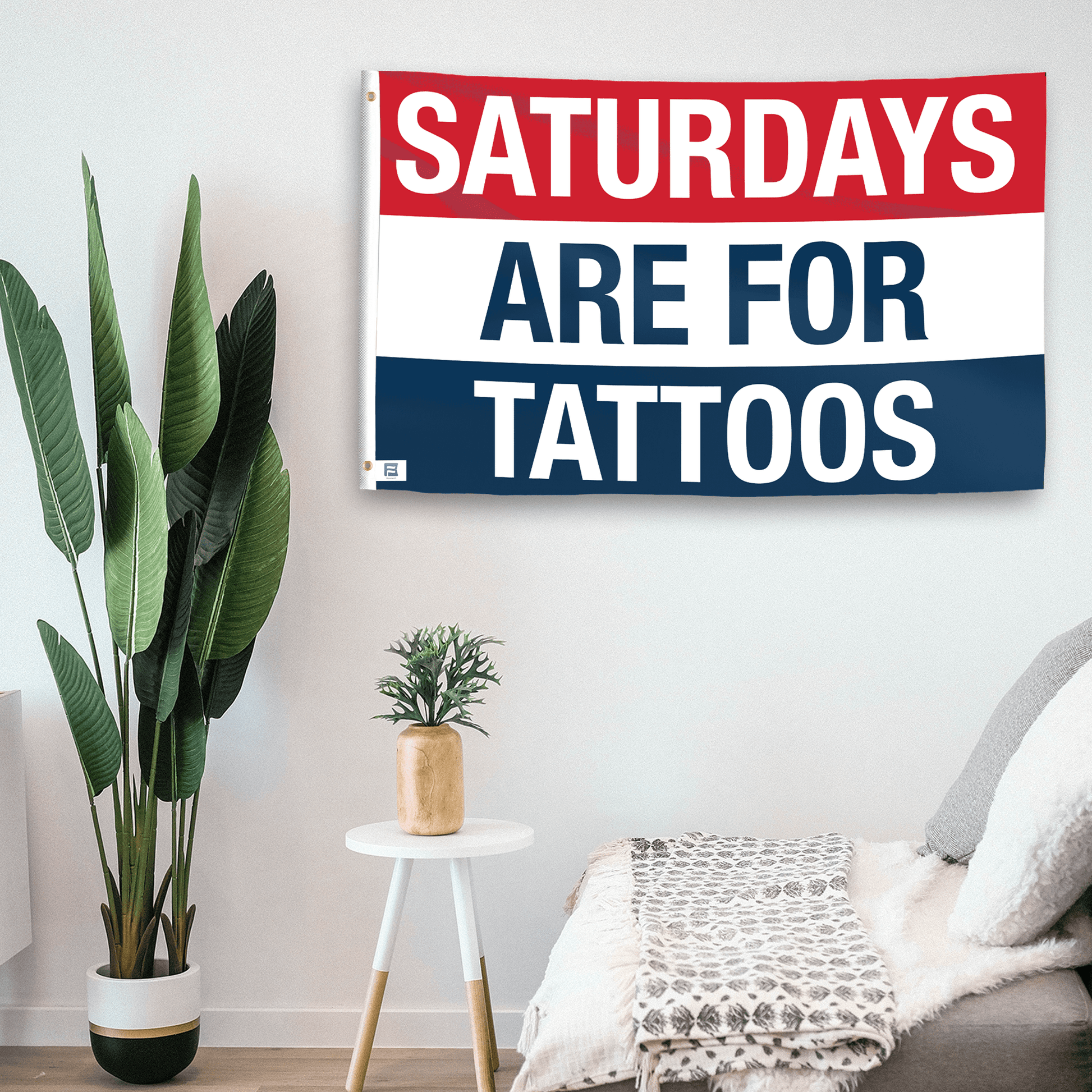 In a home setting, a flag with the saying "https://www.dropbox.com/scl/fi/fn1j4q3gdhw8bgg1y0p41/saturdays-are-for-tattoos_room.png?rlkey=2gtcisjxr8oautl6rbk4y9ip3&raw=1" is mounted on a white wall by a side table.