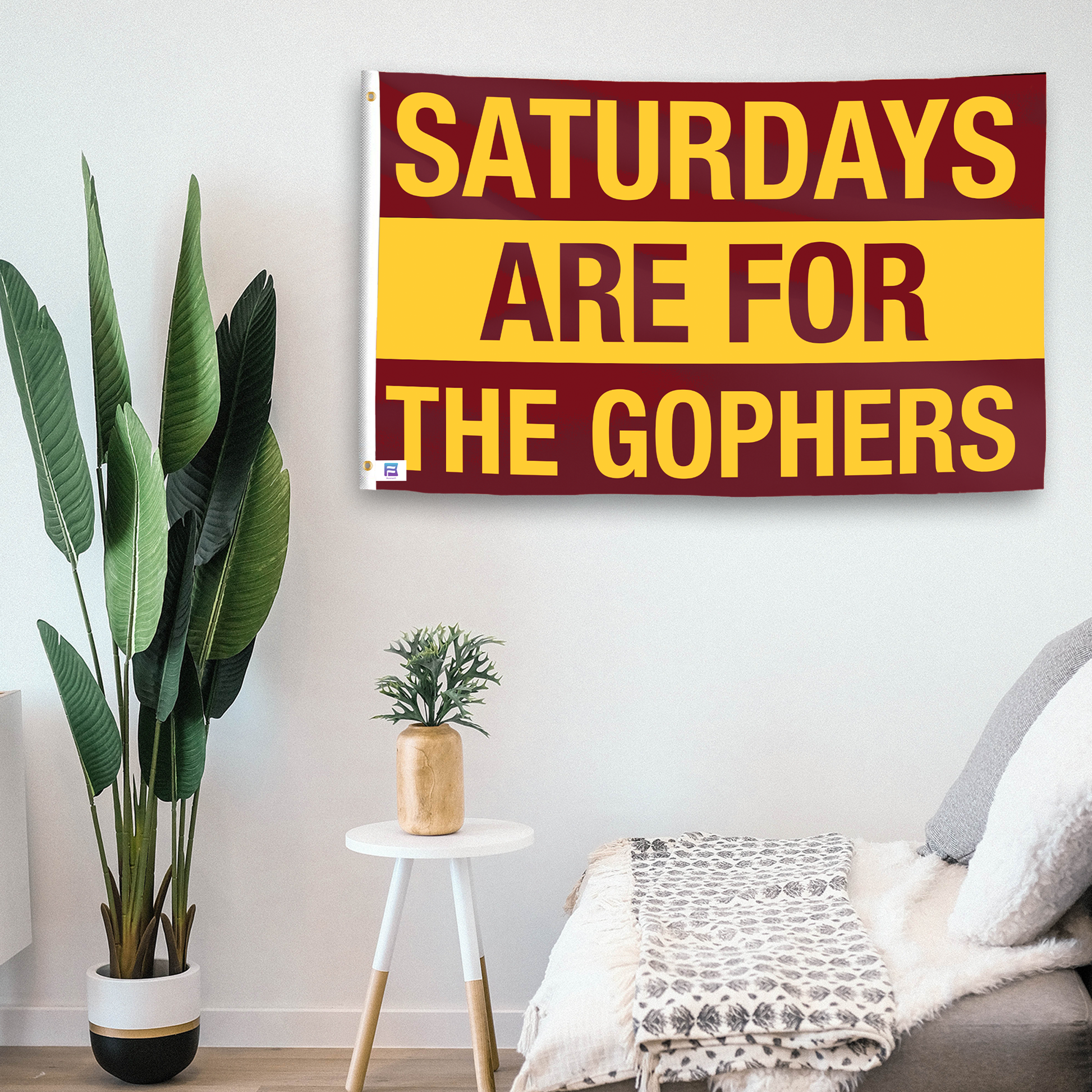 In a home setting, a flag with the saying "Saturdays Are for the Gophers" is mounted on a white wall by a side table.