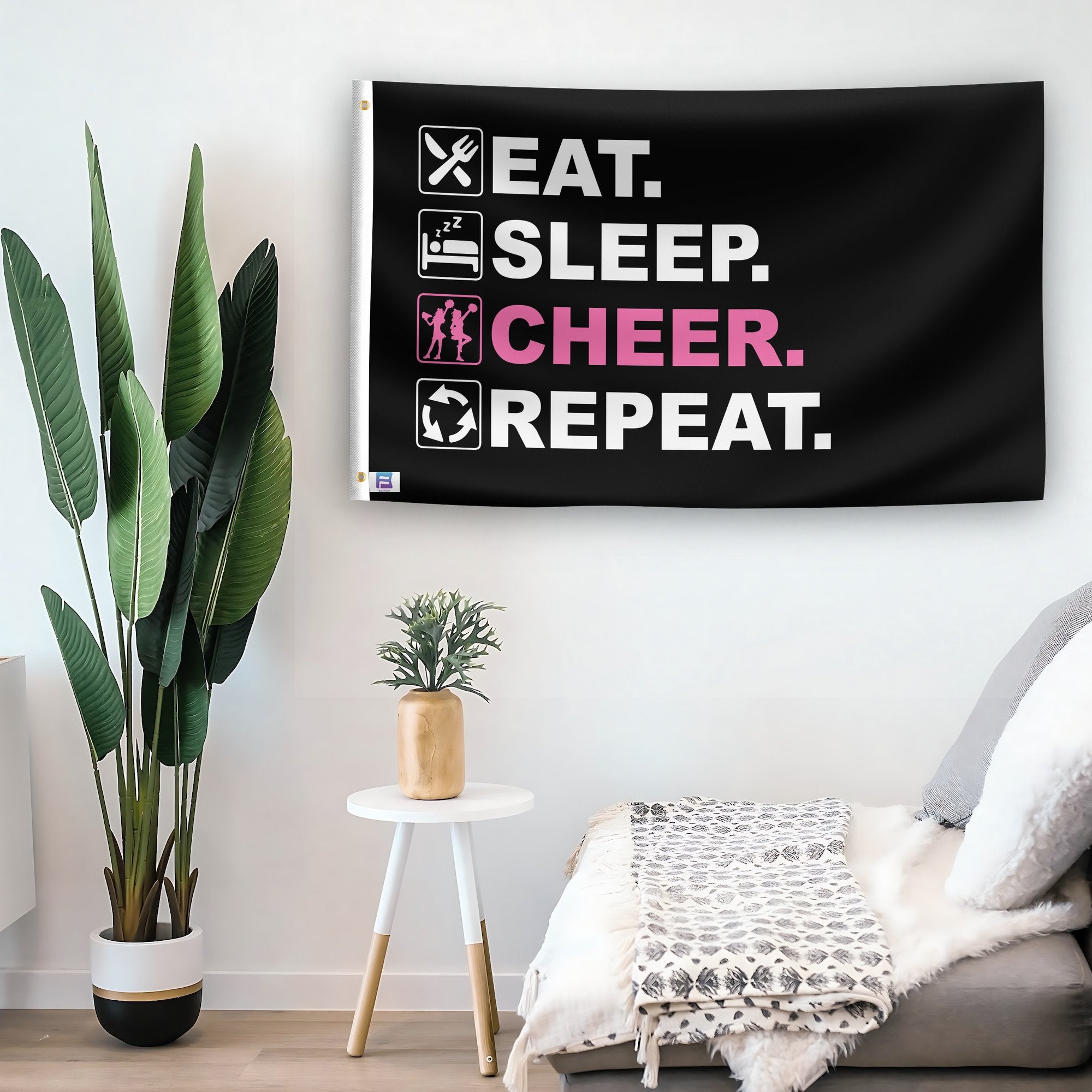 In a home setting, a flag with the saying "Eat Sleep Cheer Repeat" is mounted on a white wall by a side table.