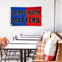 Load image into Gallery viewer, A political flag in royal blue and crimson red with a political statement on a wall in a cafe setting.
