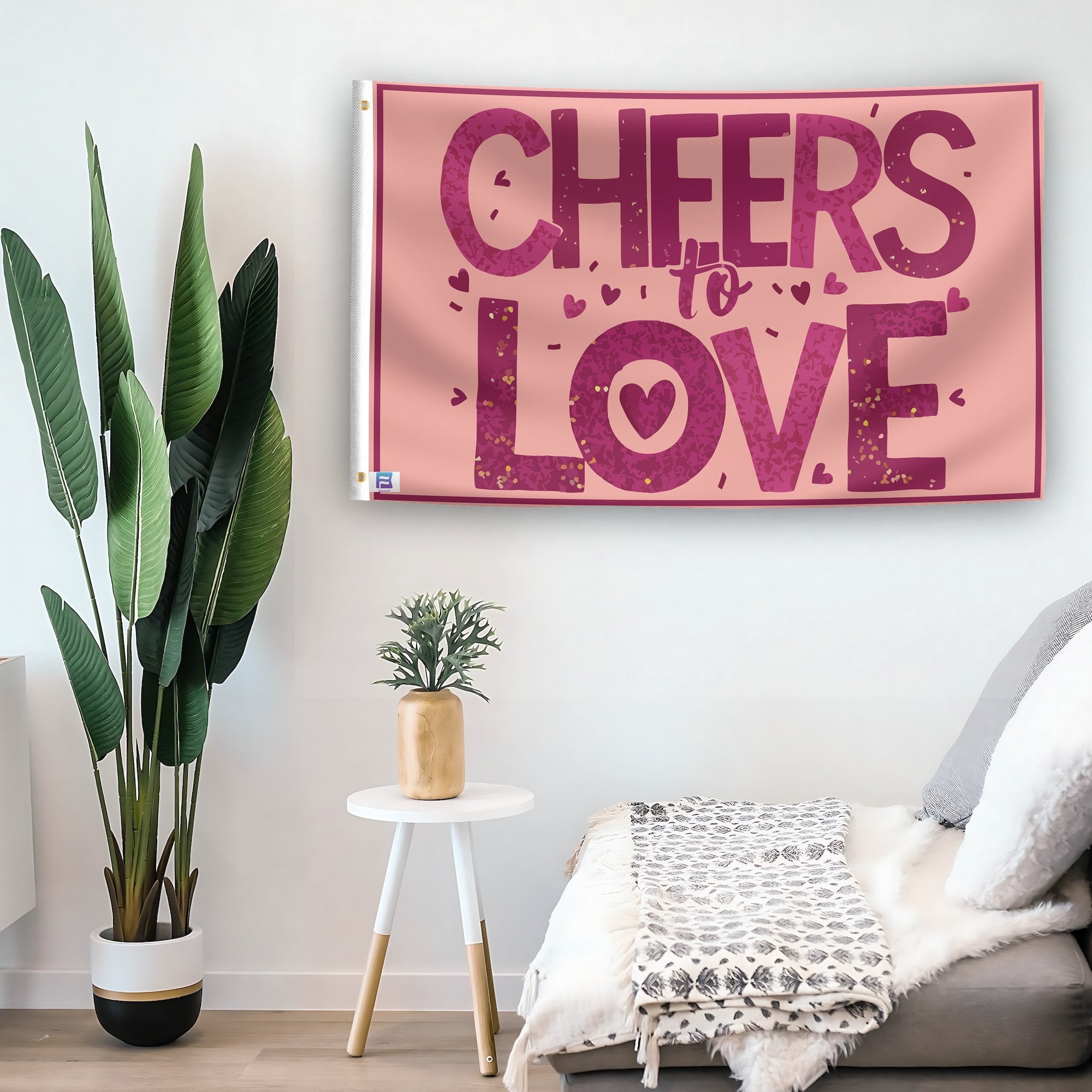 In a home setting, a flag with the saying "Cheers To Love" is mounted on a white wall by a side table.