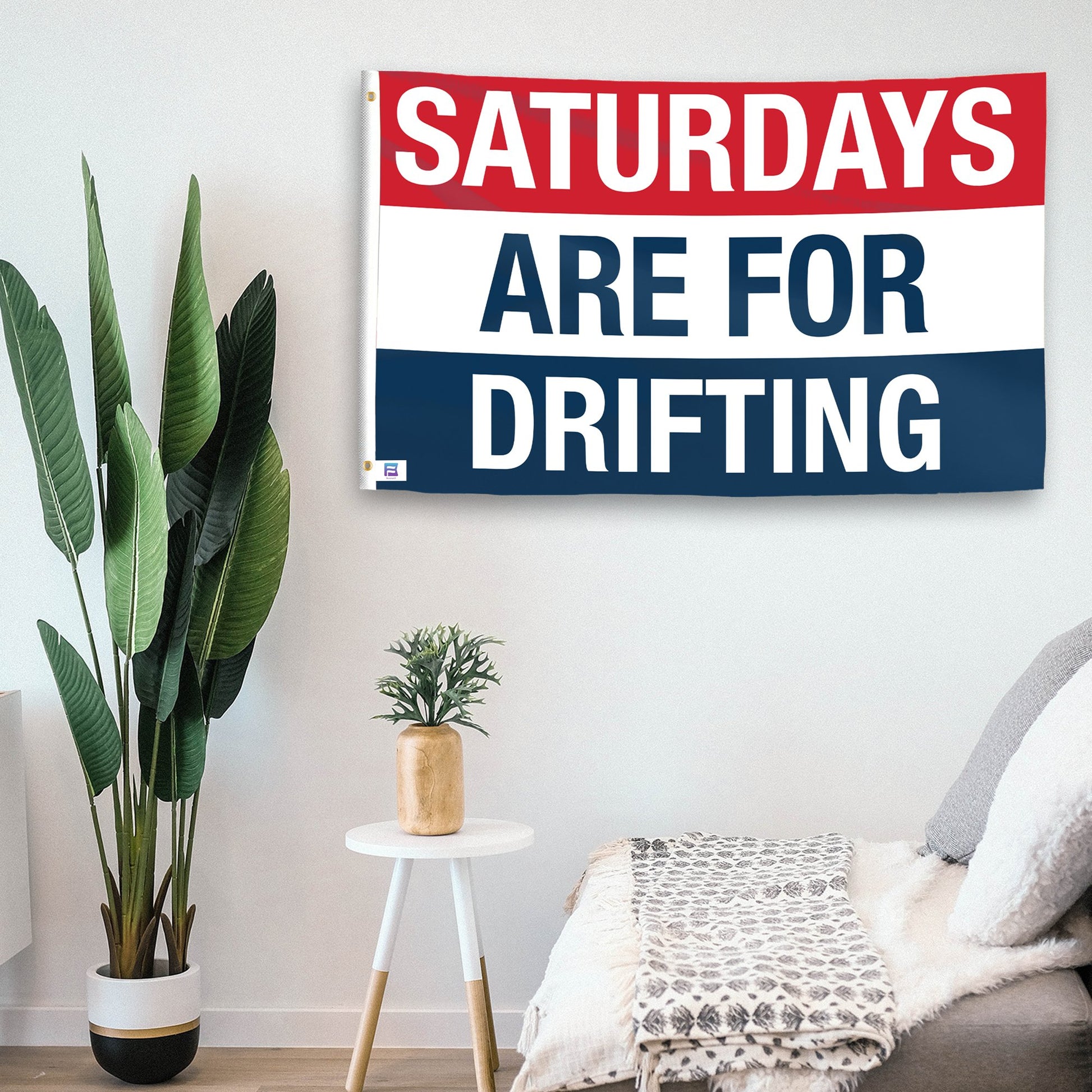 In a home setting, a flag with the saying "https://www.dropbox.com/scl/fi/rg0kahxl33ygfvvjc422q/saturdays-are-for-drifting_room.png?rlkey=sc4x8m9b73gt2a6k8m957xw9w&raw=1" is mounted on a white wall by a side table.