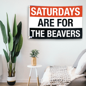 In a home setting, a flag with the saying "Saturdays Are for the Beavers" is mounted on a white wall by a side table.