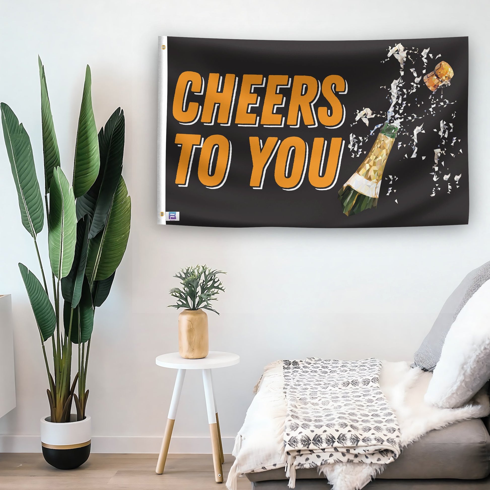 In a home setting, a flag with the saying "Cheers To You" is mounted on a white wall by a side table.