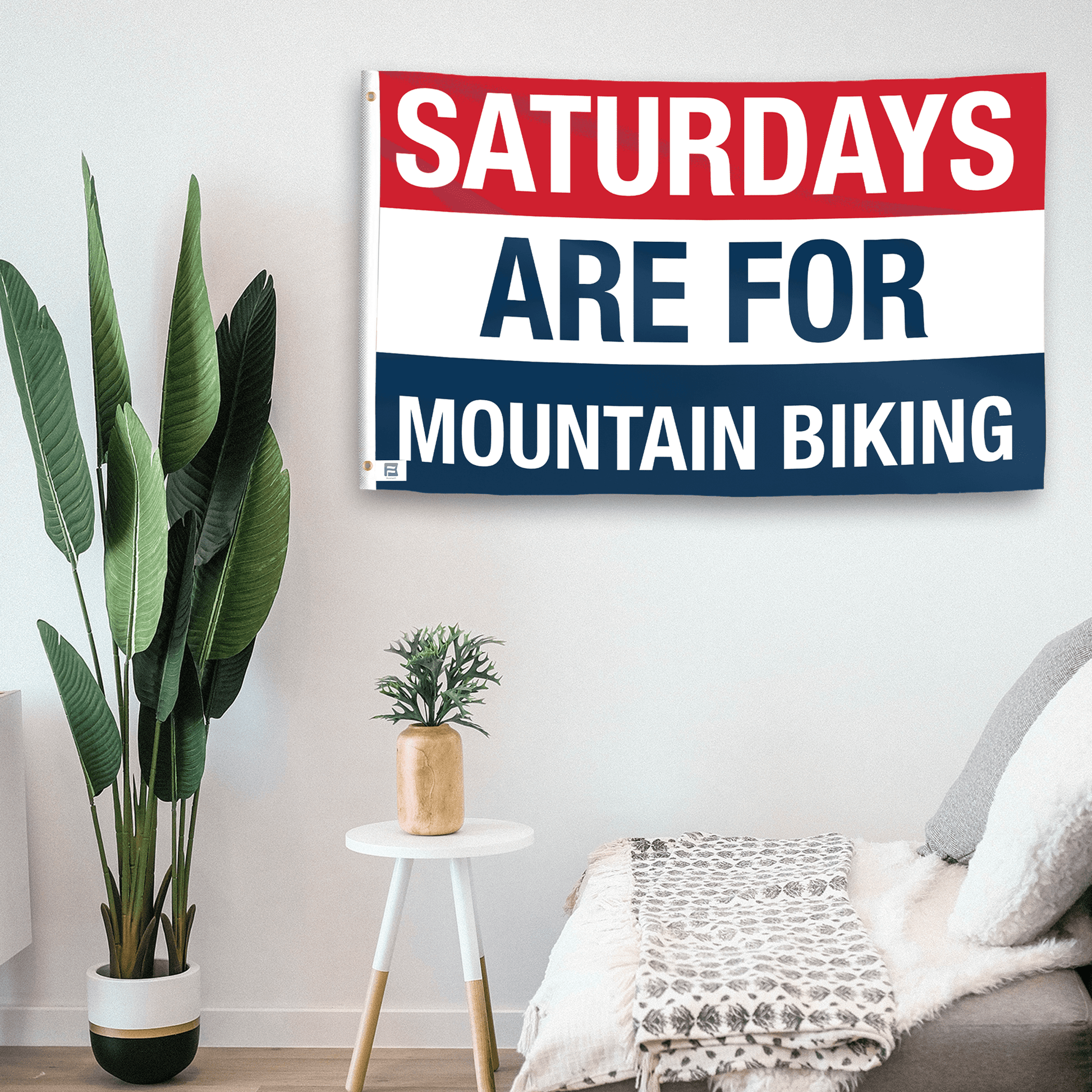 In a home setting, a flag with the saying "https://www.dropbox.com/scl/fi/2anupww4i01k8vc5gwwes/saturdays-are-for-mountain-biking_room.png?rlkey=m8qo1fohz76rd5d3z1q7j0mfz&raw=1" is mounted on a white wall by a side table.