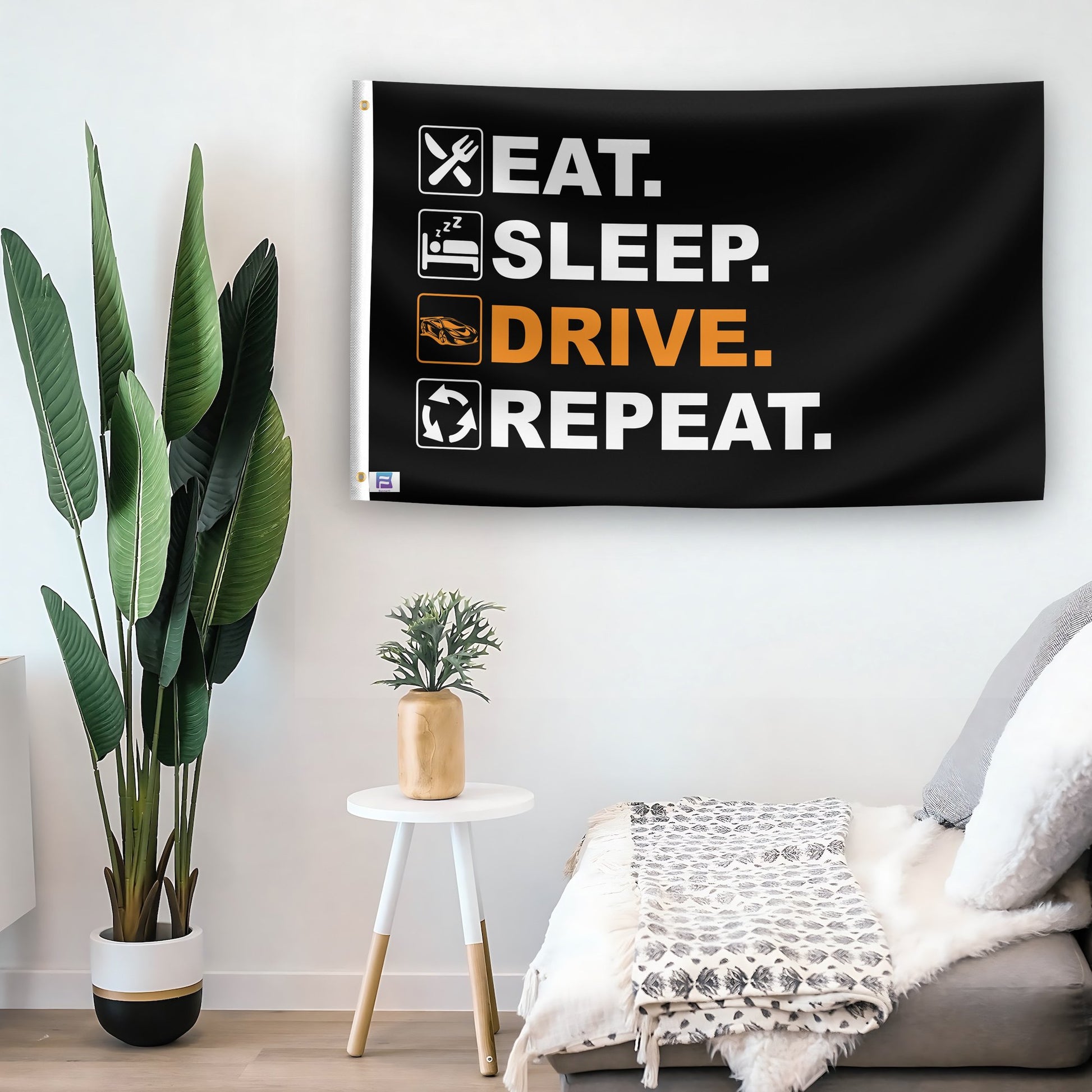 In a home setting, a flag with the saying "Eat Sleep Drive Repeat" is mounted on a white wall by a side table.