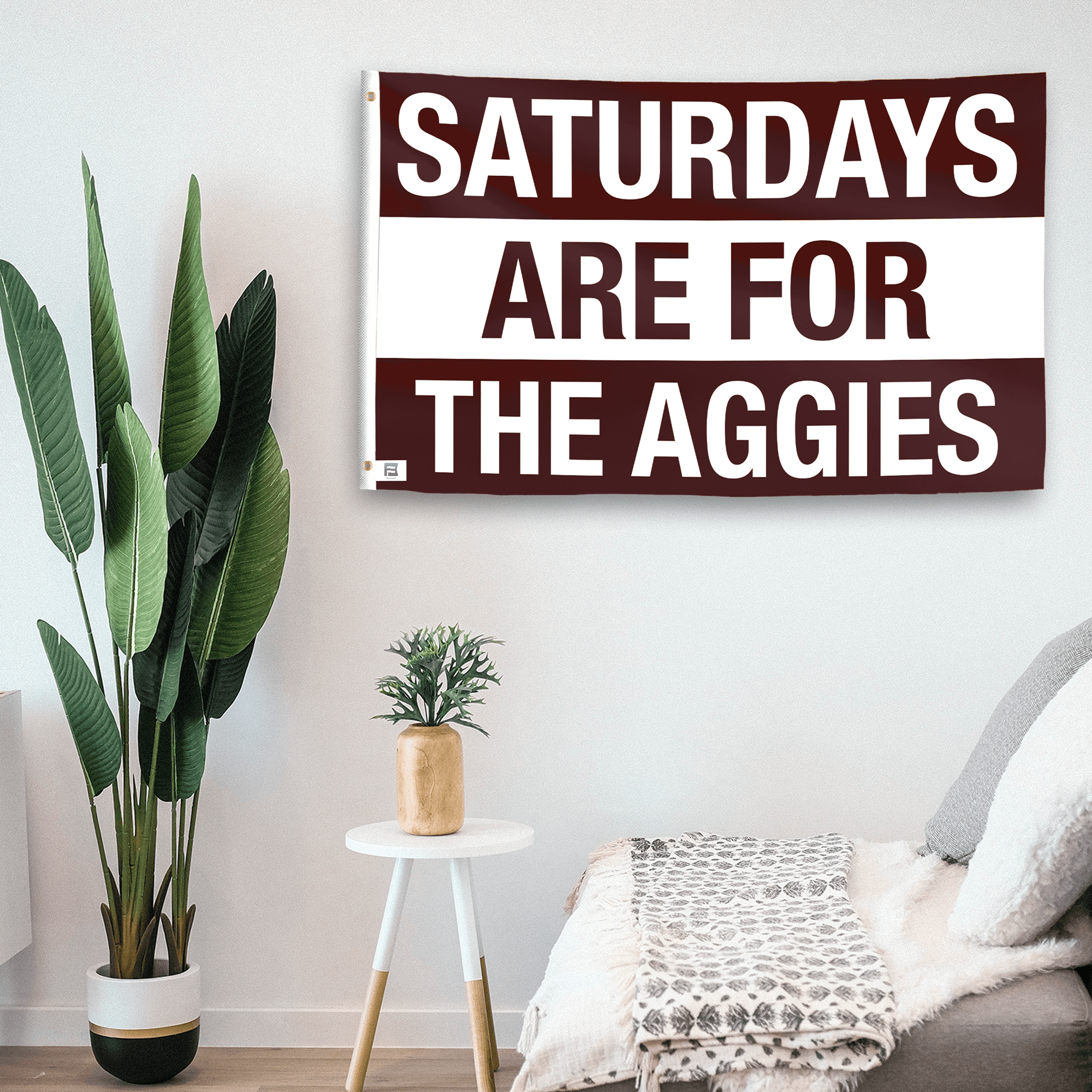 In a home setting, a flag with the saying "Saturdays Are for the Aggies" is mounted on a white wall by a side table.