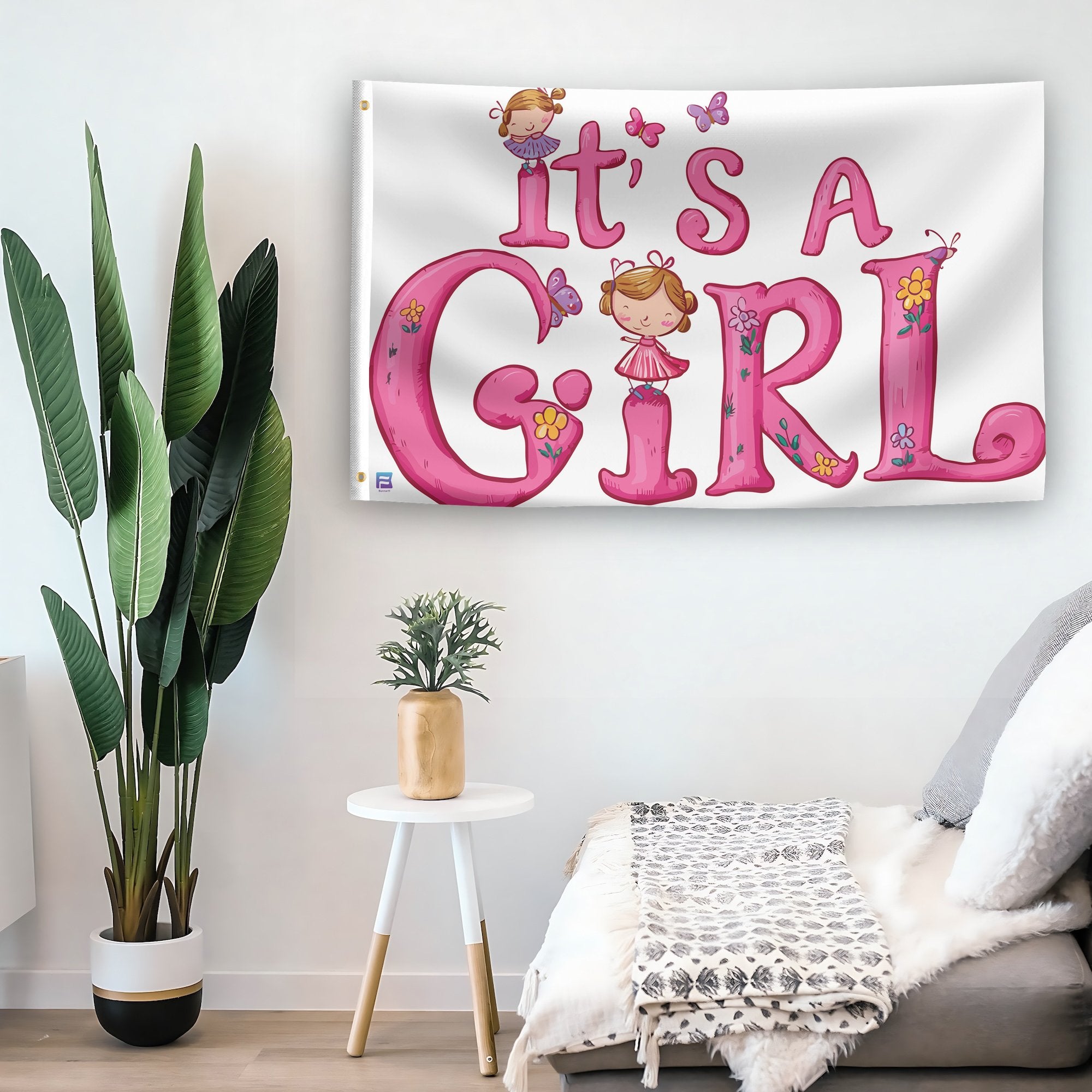 In a home setting, a flag with the saying "It's A Girl" is mounted on a white wall by a side table.