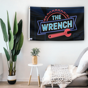 In a home setting, a flag with the saying "The Wrench" is mounted on a white wall by a side table.