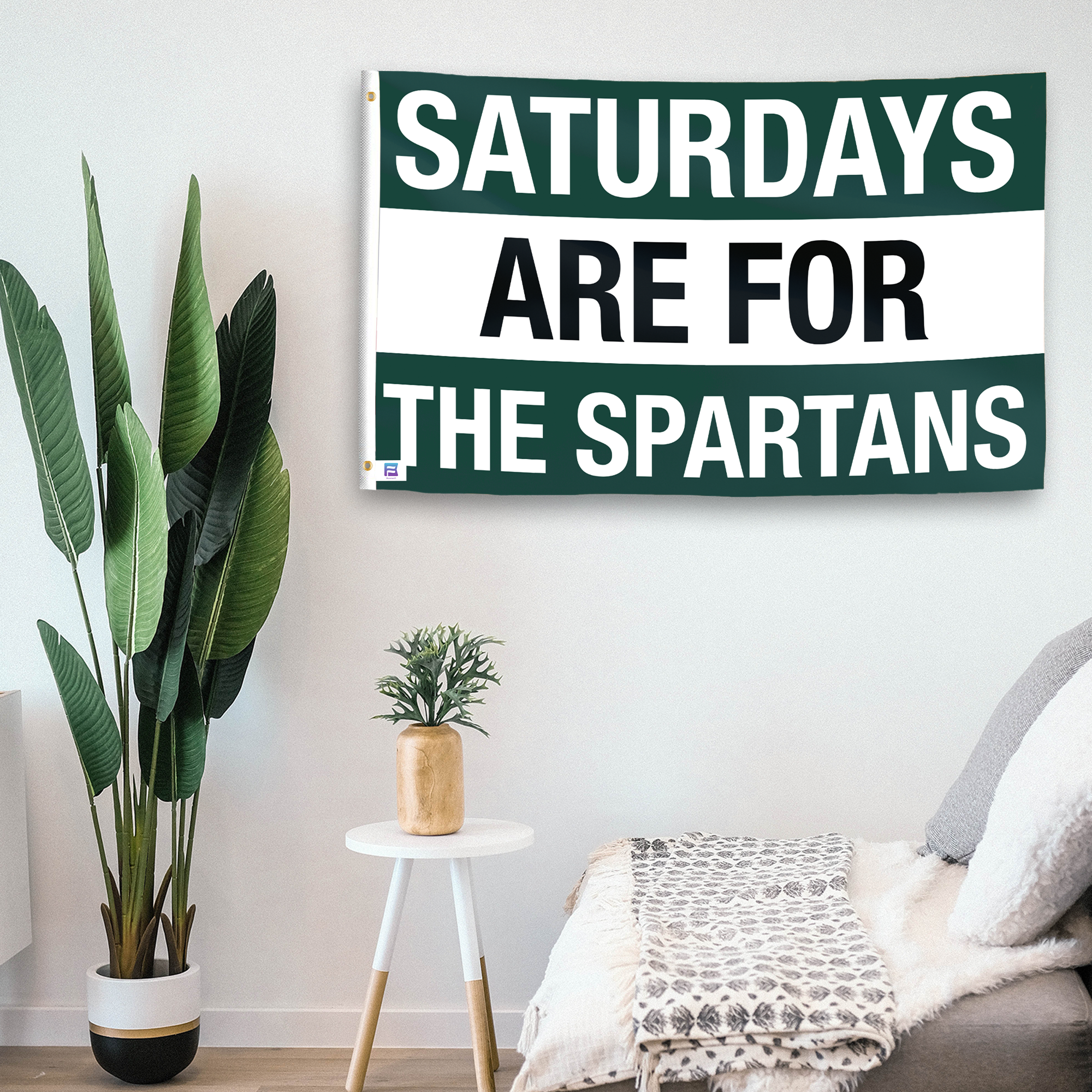 In a home setting, a flag with the saying "Saturdays Are for the Spartans" is mounted on a white wall by a side table.