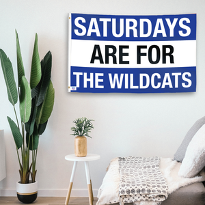 In a home setting, a flag with the saying "Saturdays Are for the Wildcats Kentucky" is mounted on a white wall by a side table.