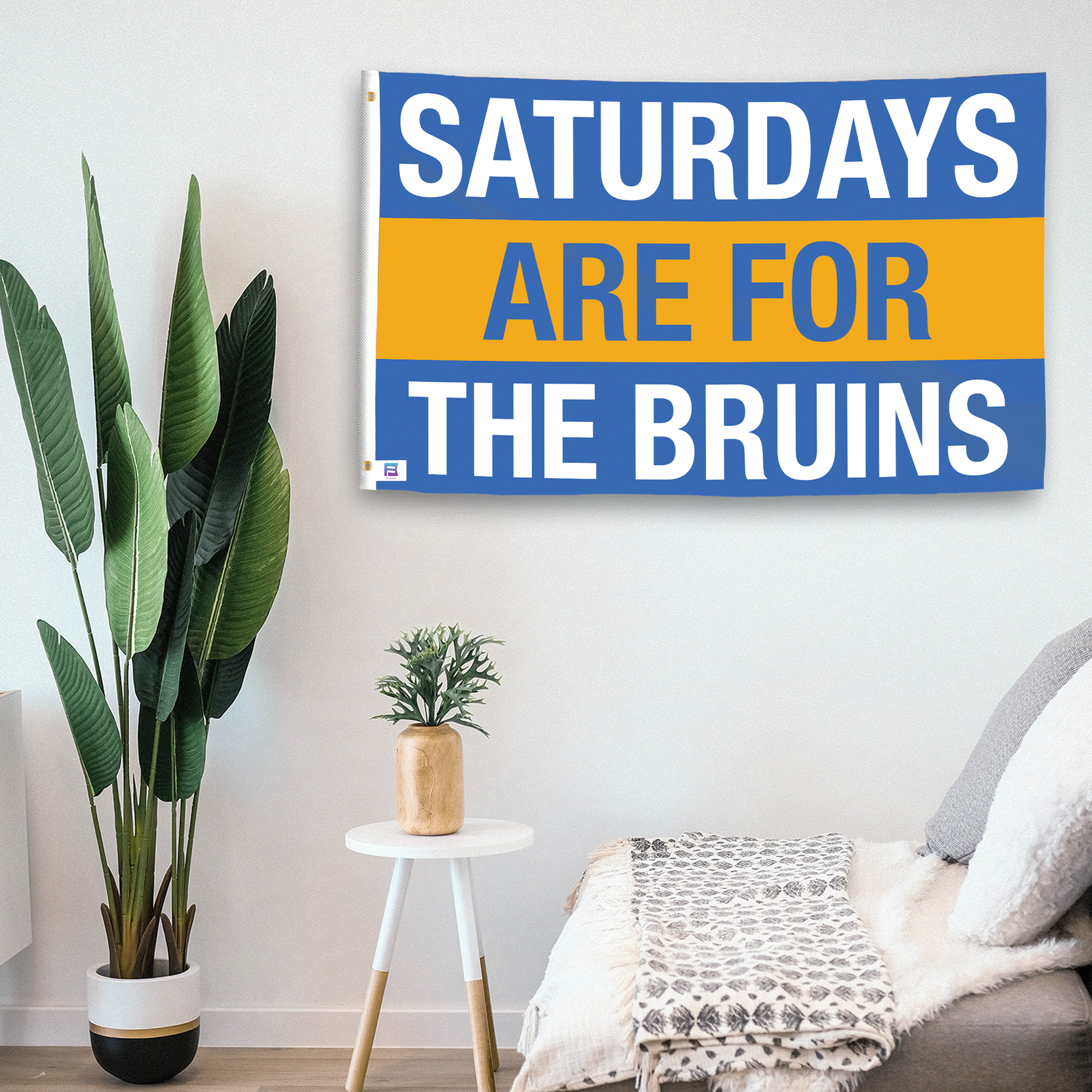 In a home setting, a flag with the saying "Saturdays Are for the Bruins" is mounted on a white wall by a side table.