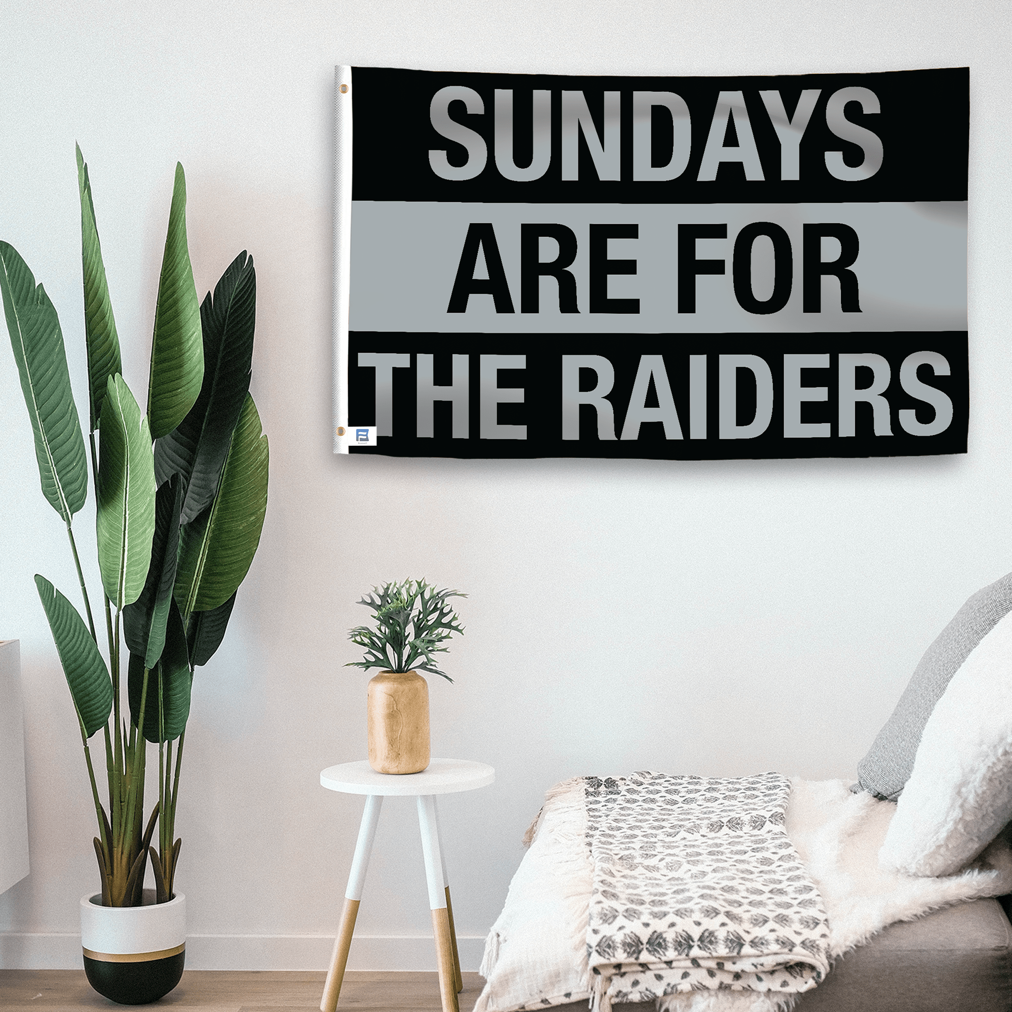 In a home setting, a flag with the saying "https://www.dropbox.com/scl/fi/s7fz7o8ms7y14l8j0tvua/sundays-are-for-the-raiders_room.png?rlkey=zm37xpbfgi0ybme48r2r21cst&raw=1" is mounted on a white wall by a side table.