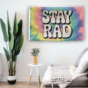 In a home setting, a flag with the saying "Stay Rad" is mounted on a white wall by a side table.