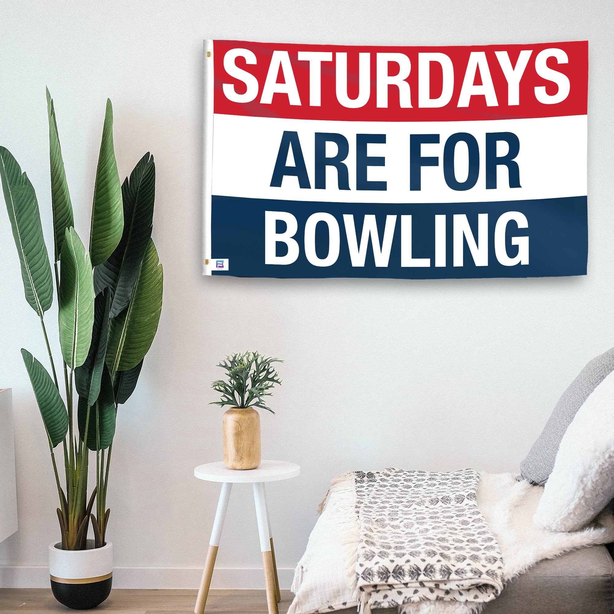 In a home setting, a flag with the saying "https://www.dropbox.com/scl/fi/8wkaest3npm1p5foc8bci/saturdays-are-for-bowling_room.png?rlkey=ef72opyw6pxw8vyoeev687n87&raw=1" is mounted on a white wall by a side table.