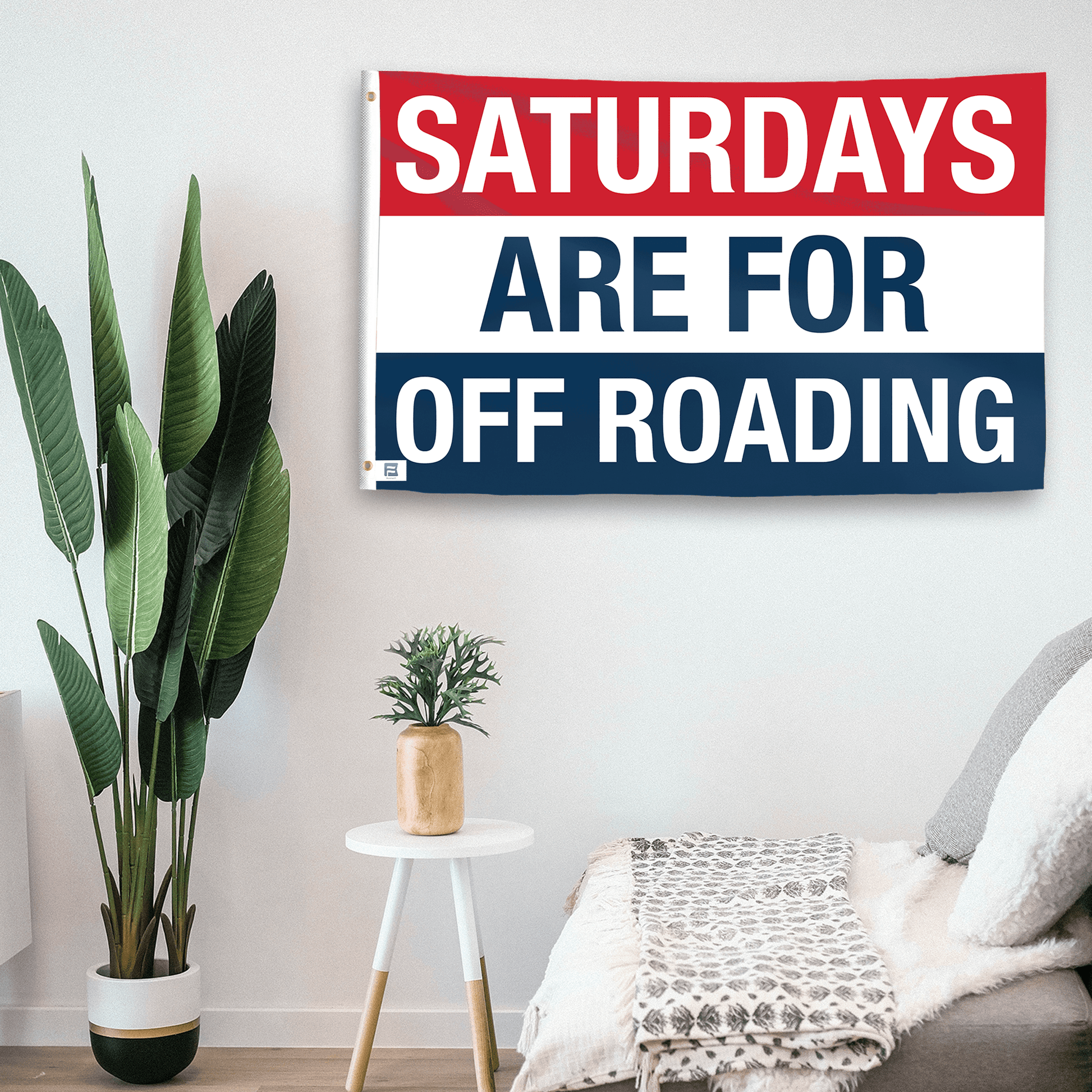 In a home setting, a flag with the saying "https://www.dropbox.com/scl/fi/h0bs7lphy5aw9kf8qzy4k/saturdays-are-for-off-roading_room.png?rlkey=ro838awrbrjo0mpek50sa7ffw&raw=1" is mounted on a white wall by a side table.