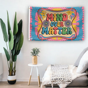 In a home setting, a flag with the saying "Mind Over Matter" is mounted on a white wall by a side table.