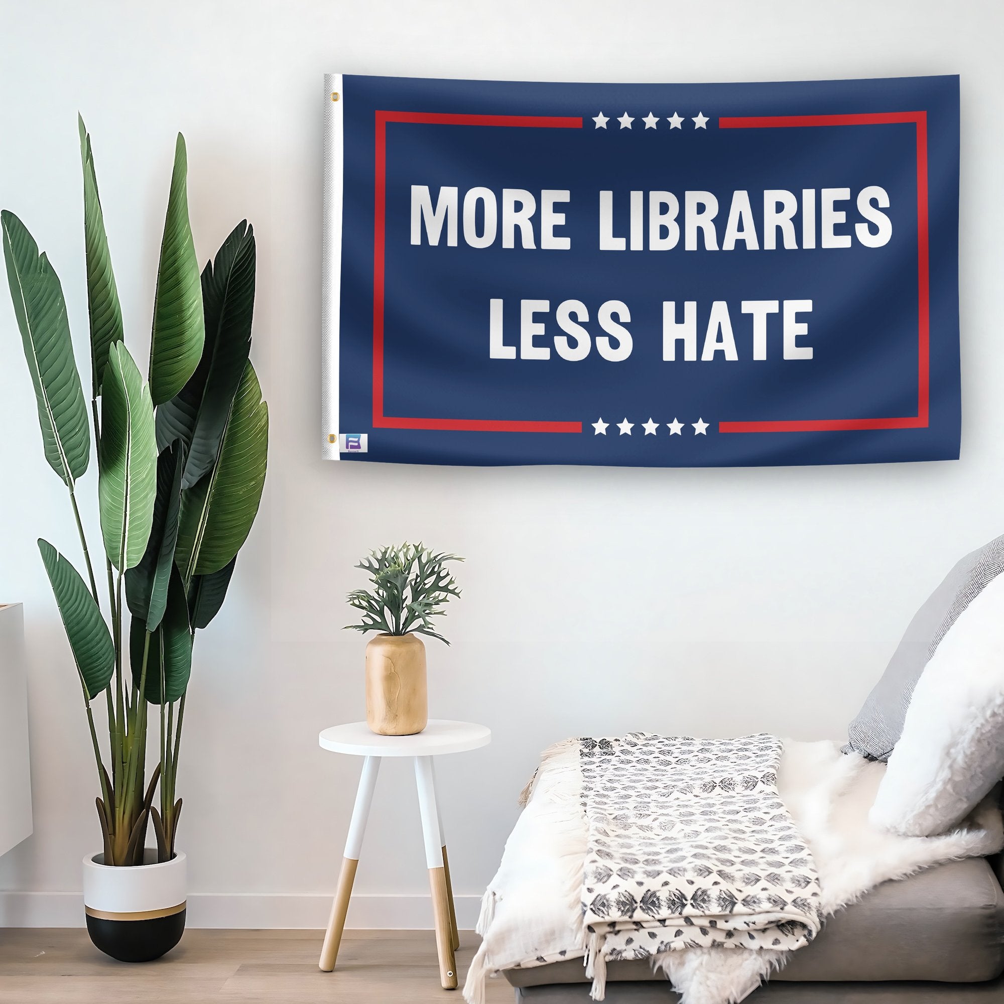 In a home setting, a flag with the saying "More Libraries Less Hate Political" is mounted on a white wall by a side table.