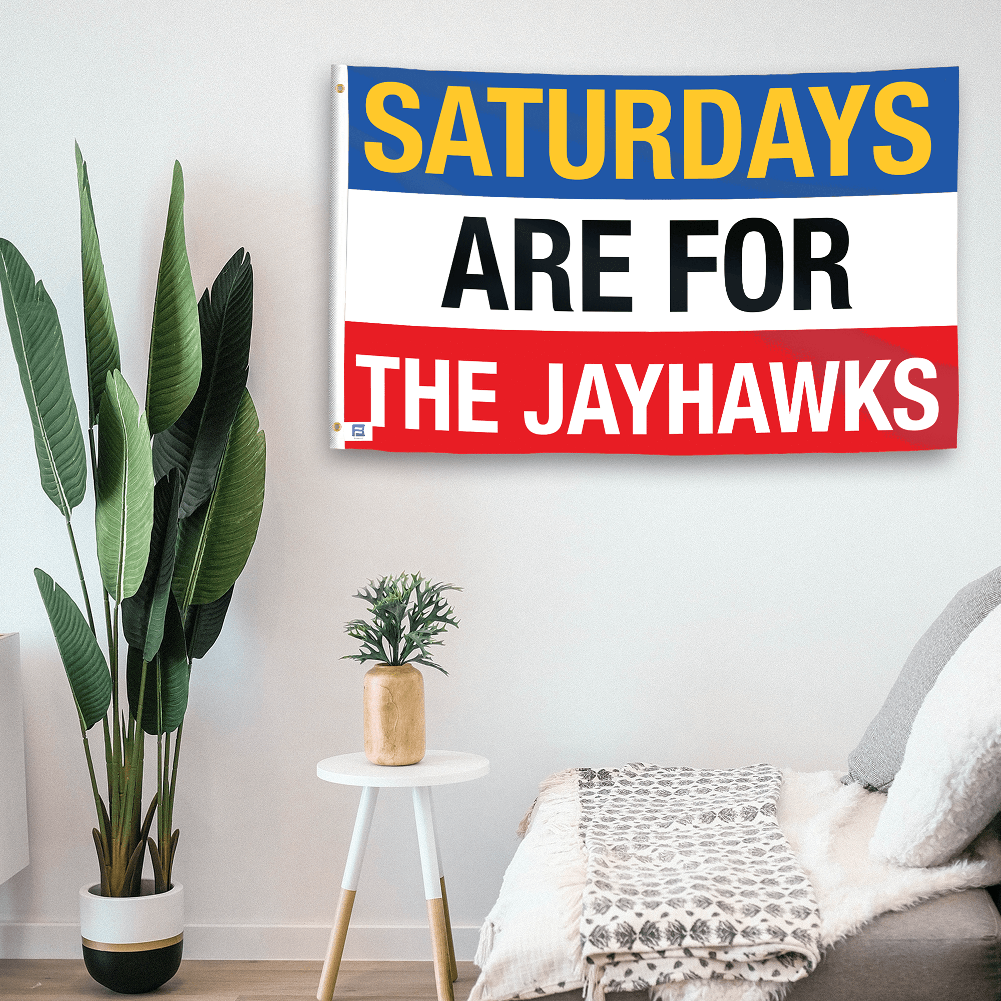 In a home setting, a flag with the saying "Saturdays Are for the Jayhawks" is mounted on a white wall by a side table.