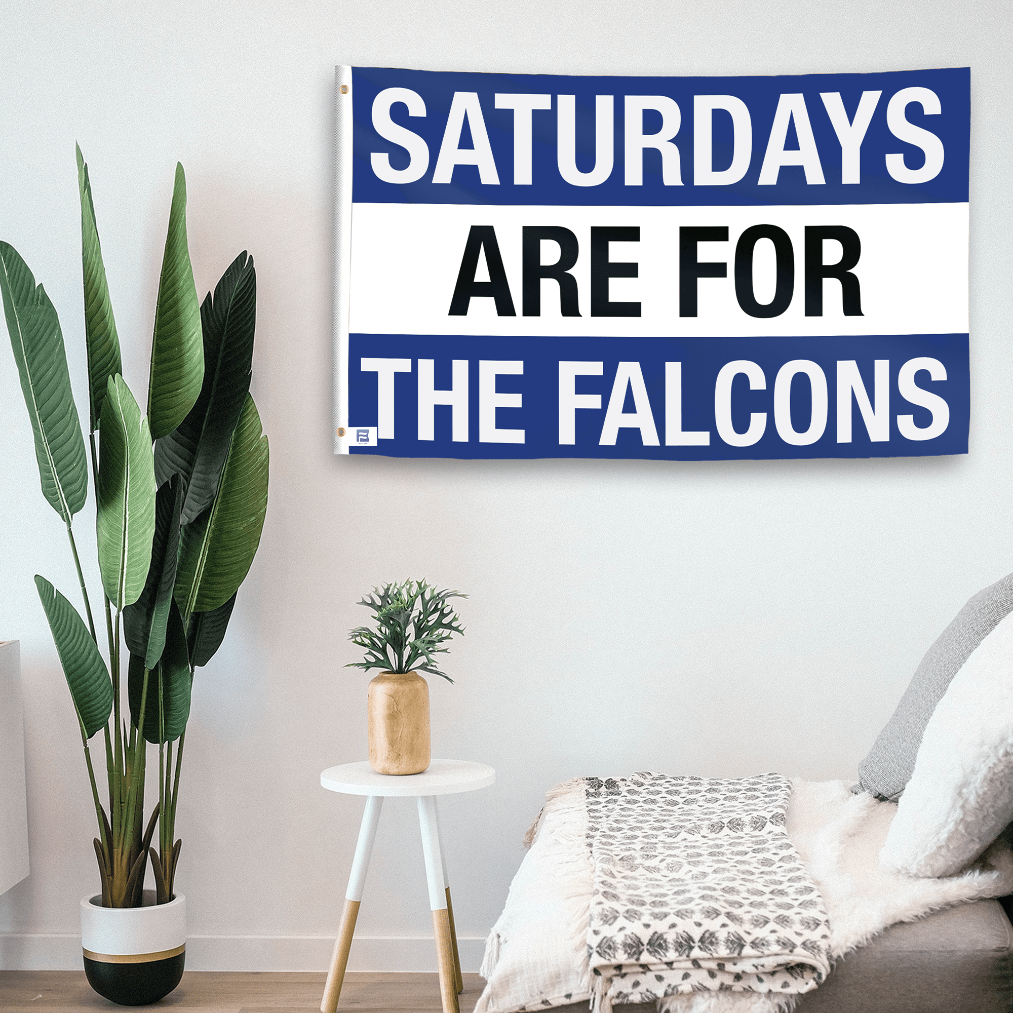 In a home setting, a flag with the saying "Saturdays Are for the Falcons" is mounted on a white wall by a side table.