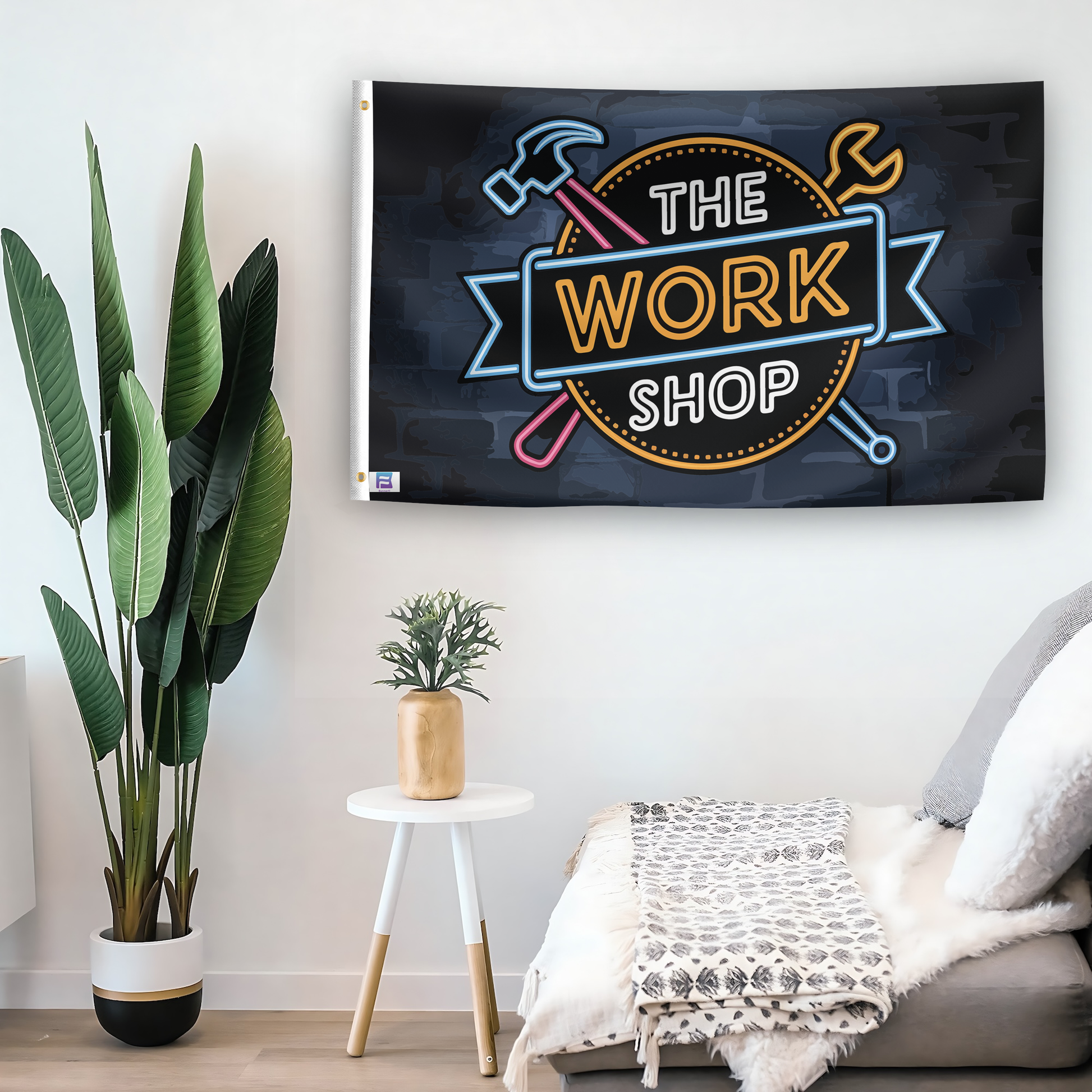 In a home setting, a flag with the saying "The Work Shop" is mounted on a white wall by a side table.