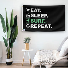 Load image into Gallery viewer, In a home setting, a flag with the saying &quot;Eat Sleep Surf Repeat&quot; is mounted on a white wall by a side table.
