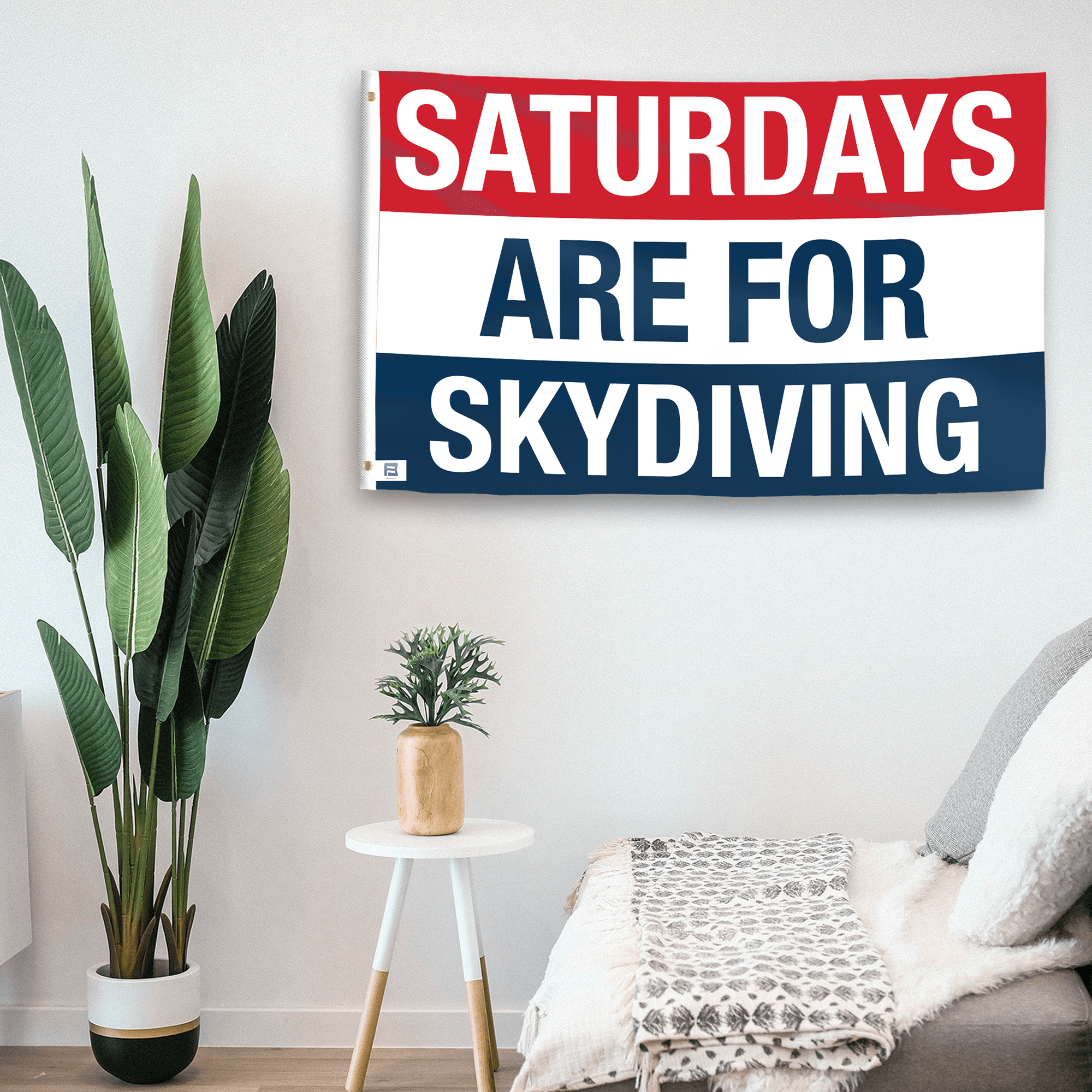 In a home setting, a flag with the saying "https://www.dropbox.com/scl/fi/5bxl2pdhcm4ib3ypkozun/saturdays-are-for-skydiving_room.png?rlkey=pcu3nxpf6szgcvso28iaab1tb&raw=1" is mounted on a white wall by a side table.