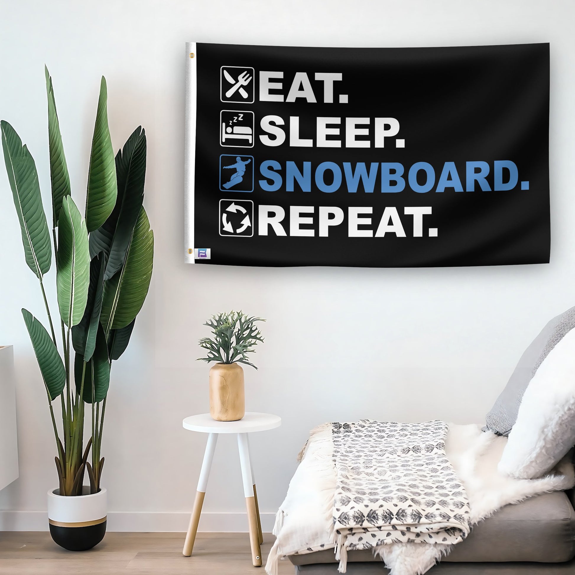 In a home setting, a flag with the saying "Eat Sleep Snowboard Repeat" is mounted on a white wall by a side table.