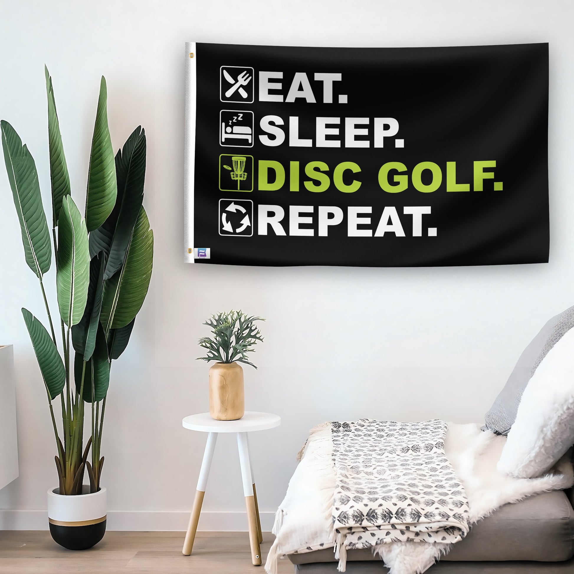 In a home setting, a flag with the saying "Eat Sleep Disc Golf Repeat" is mounted on a white wall by a side table.