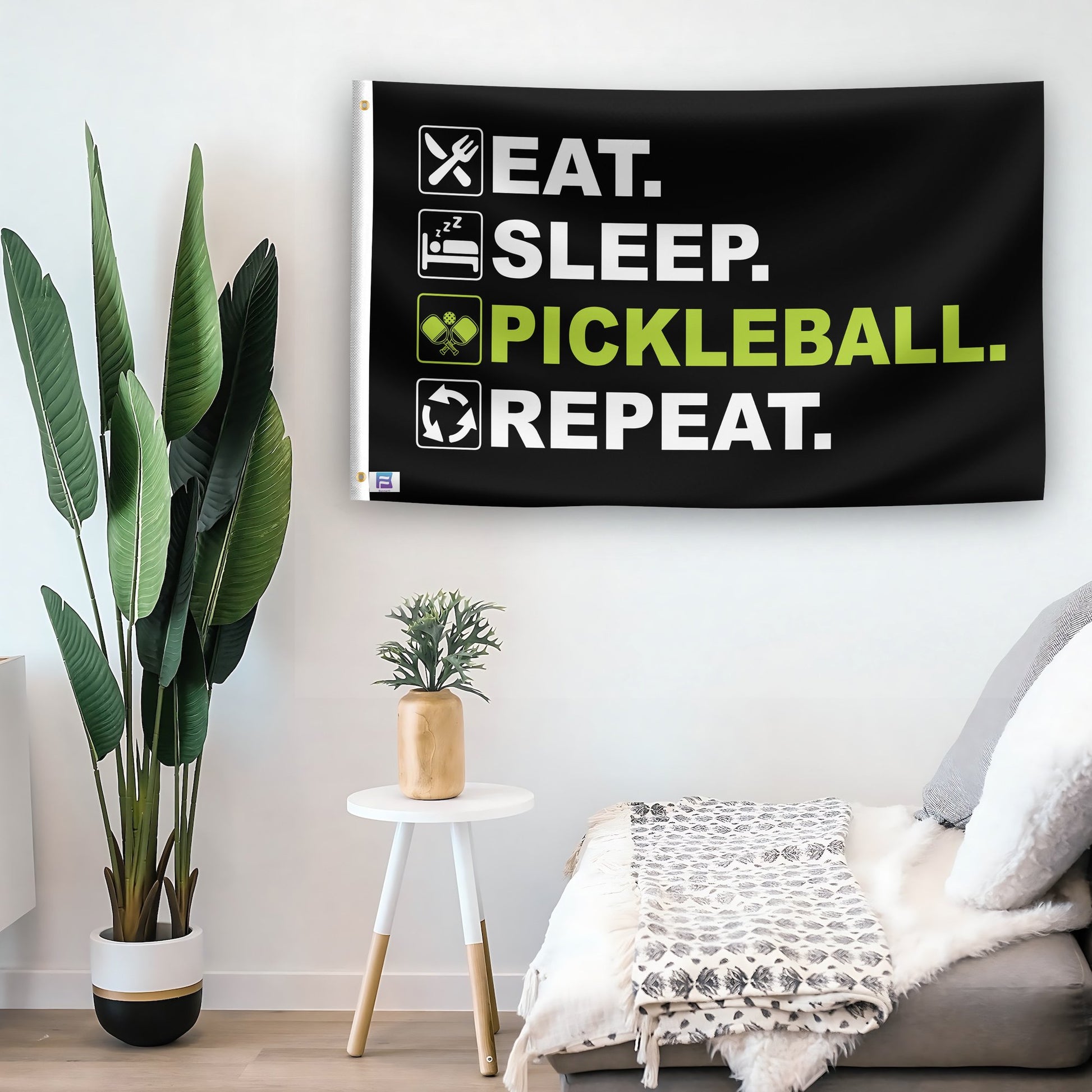 In a home setting, a flag with the saying "Eat Sleep Pickleball Repeat" is mounted on a white wall by a side table.