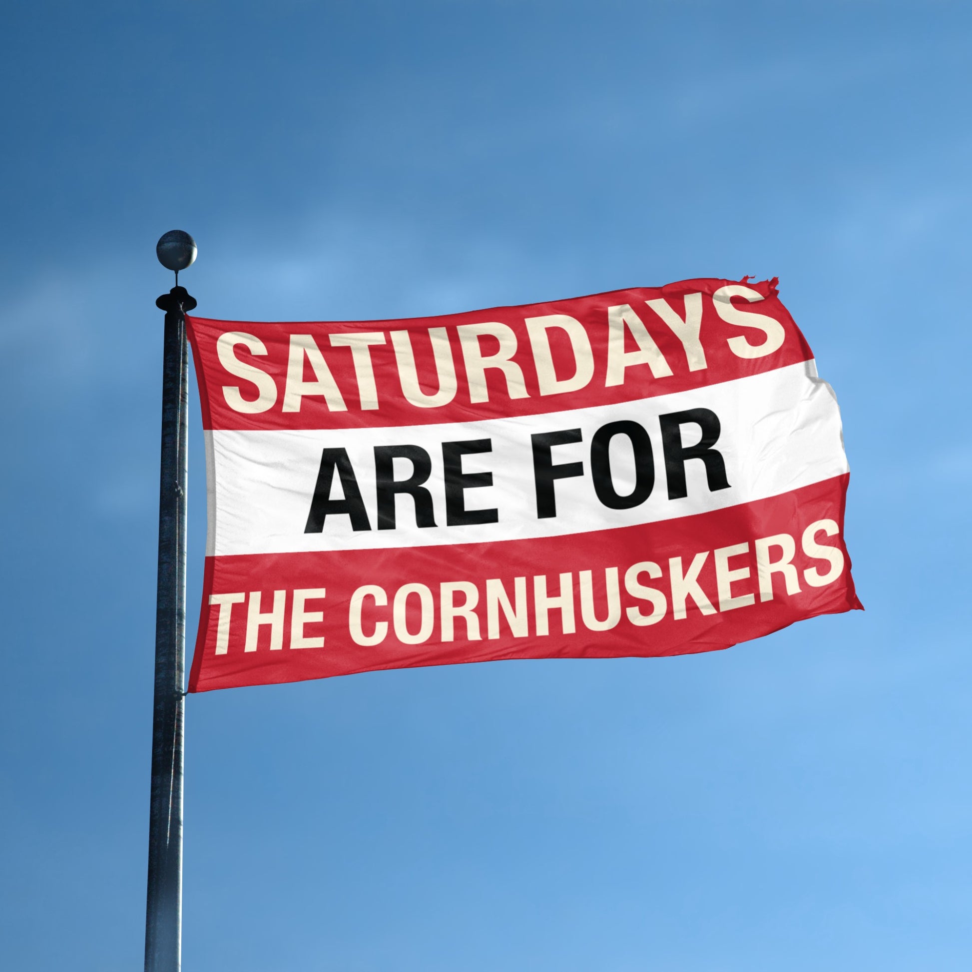Flag with text 'Saturdays are for the Cornhuskers' on a flagpole against a blue sky.