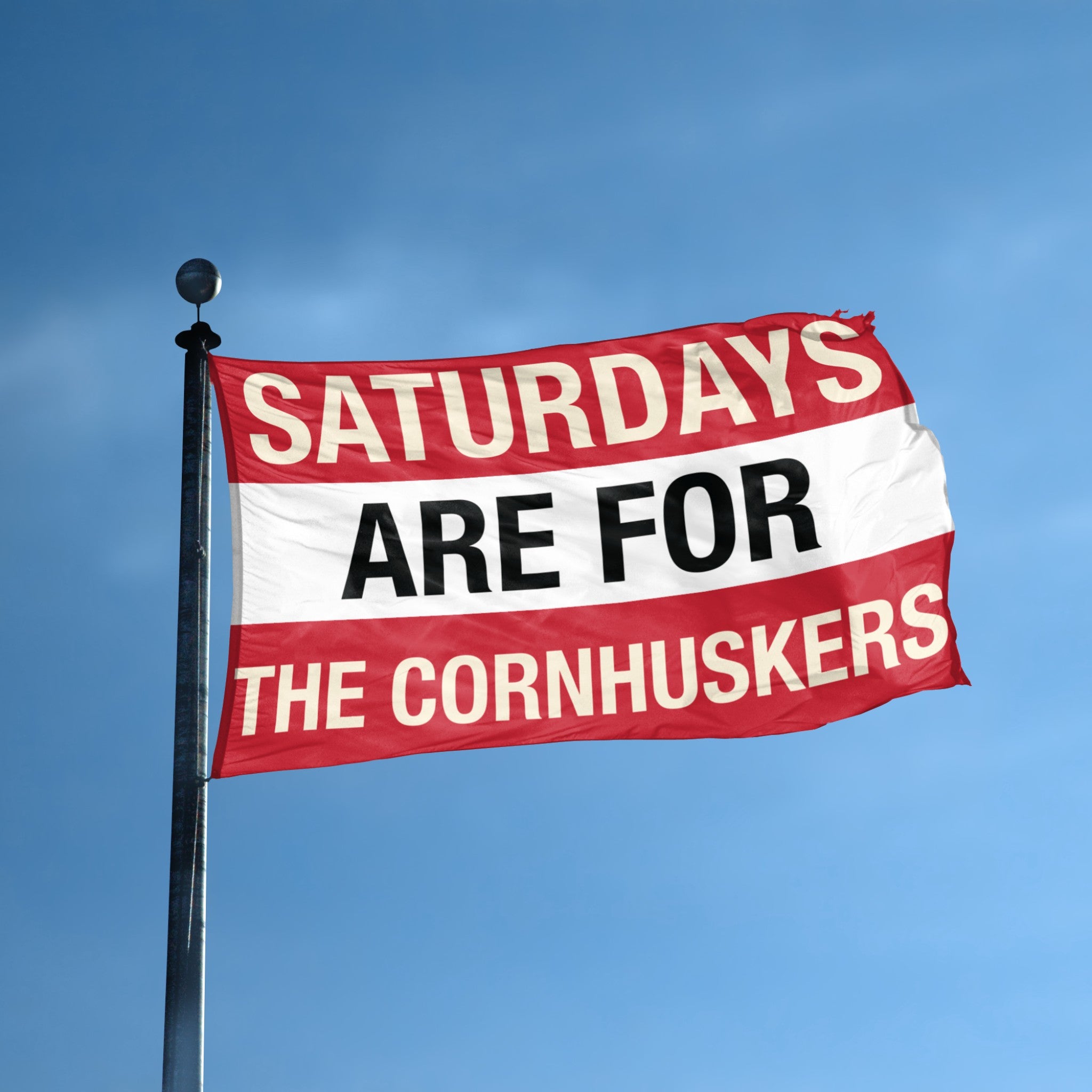 Flag with text 'Saturdays are for the Cornhuskers' on a flagpole against a blue sky.